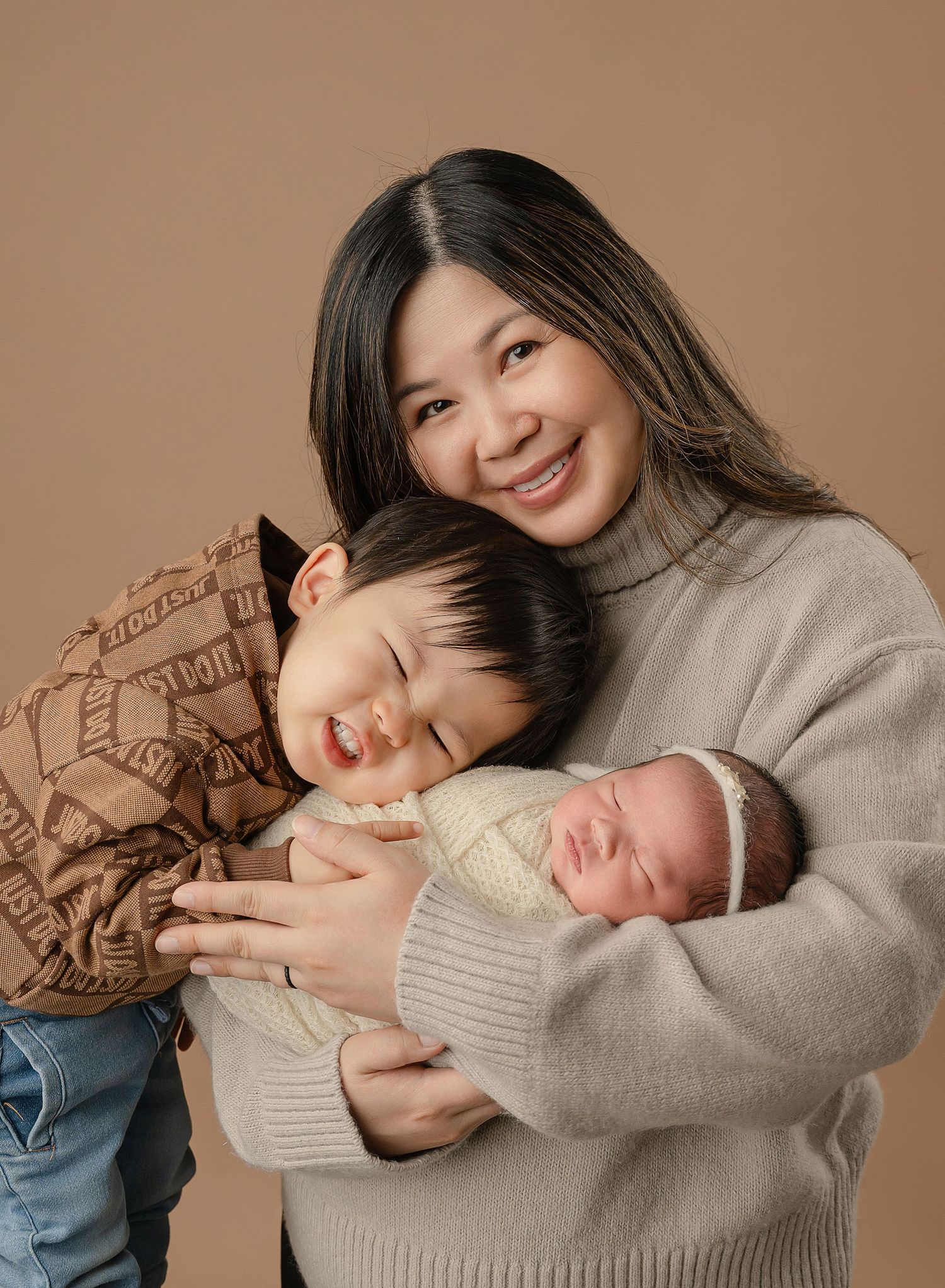 A tender family moment showing a parent in a beige sweater holding two children against a warm brown backdrop.