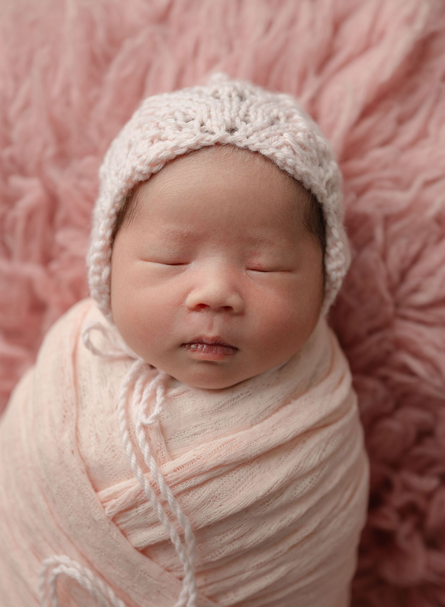 Sleeping newborn baby wrapped in soft pink swaddle wearing knitted bonnet on pink furry backdrop.