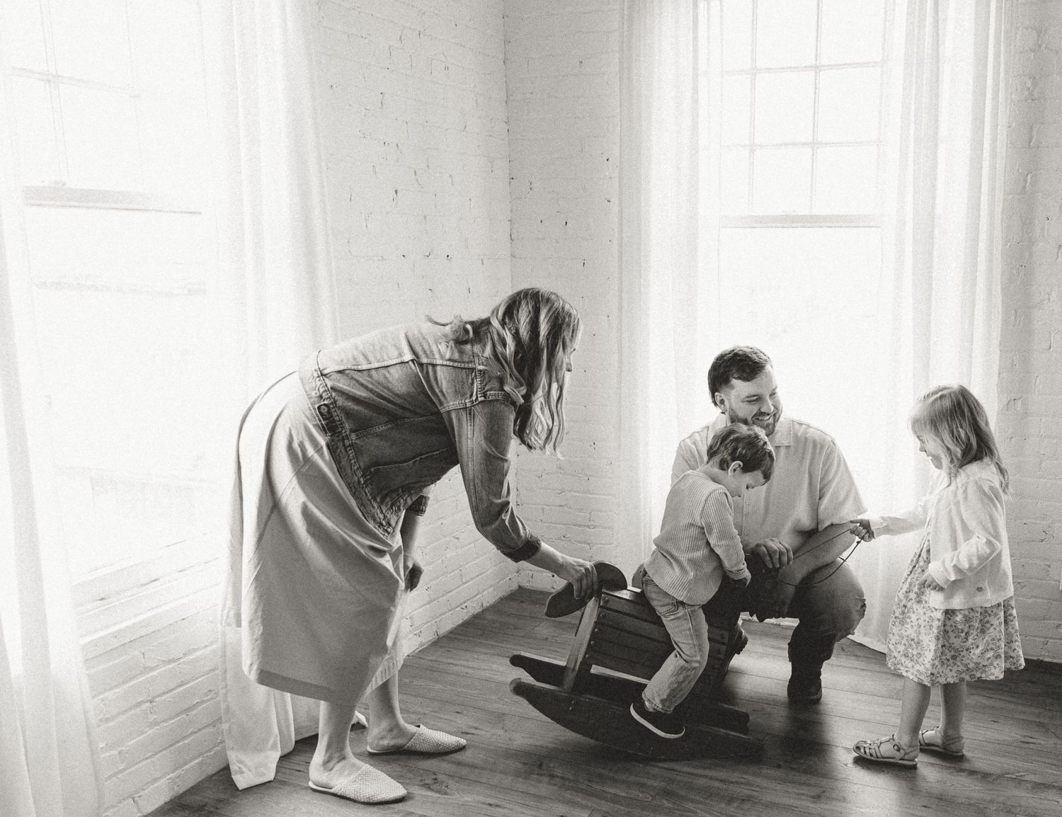 Black and white photo of family playing with rocking horse in minimalist room.