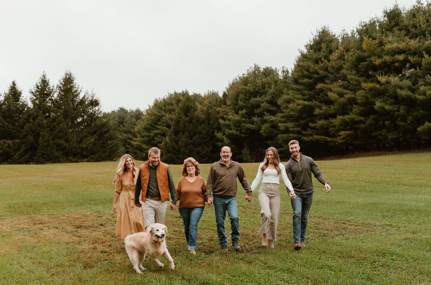 Wide landscape shot of a group walking across a green field with evergreen trees behind.