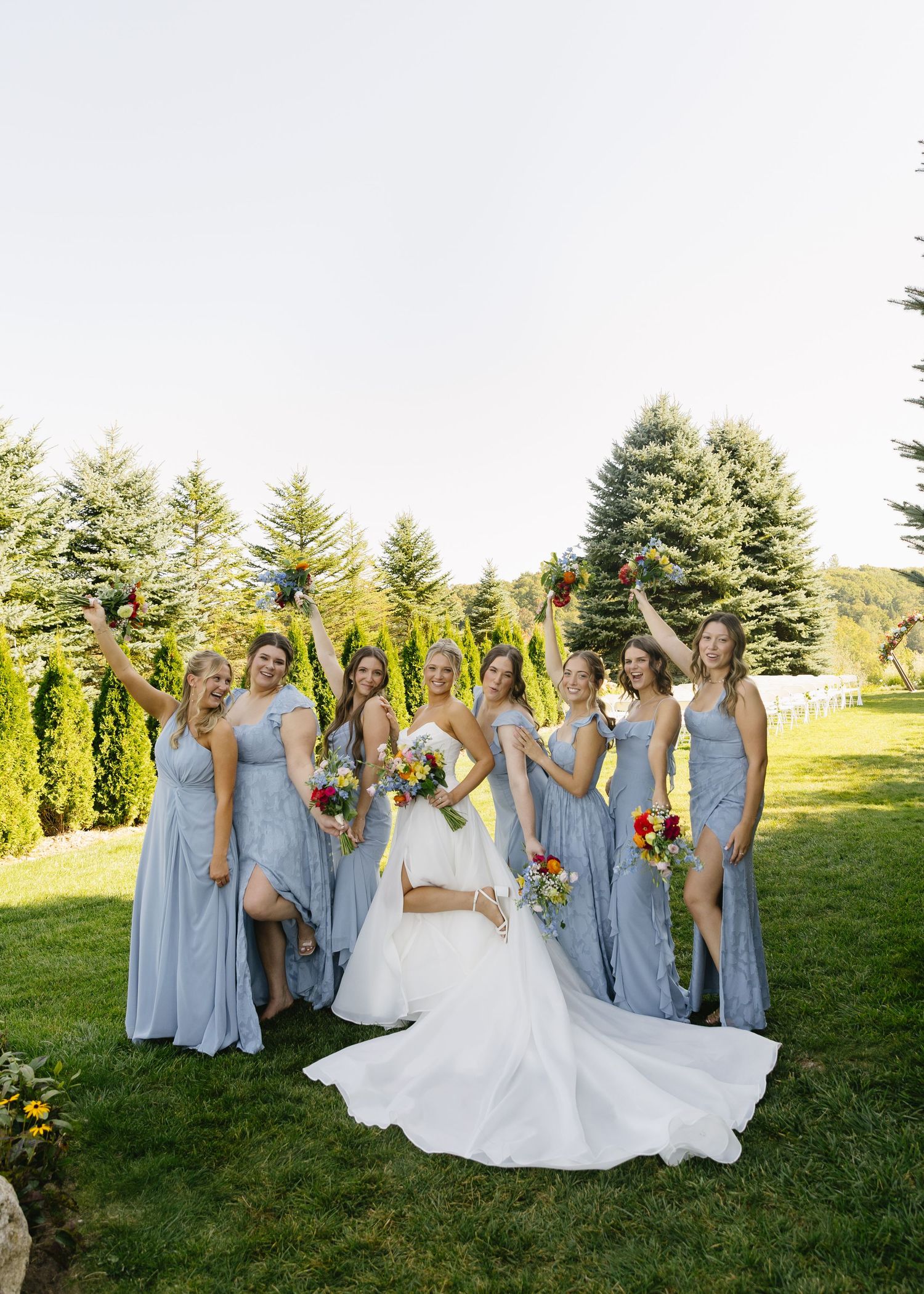 Wedding party in light blue dresses celebrates on sunny lawn with evergreens.