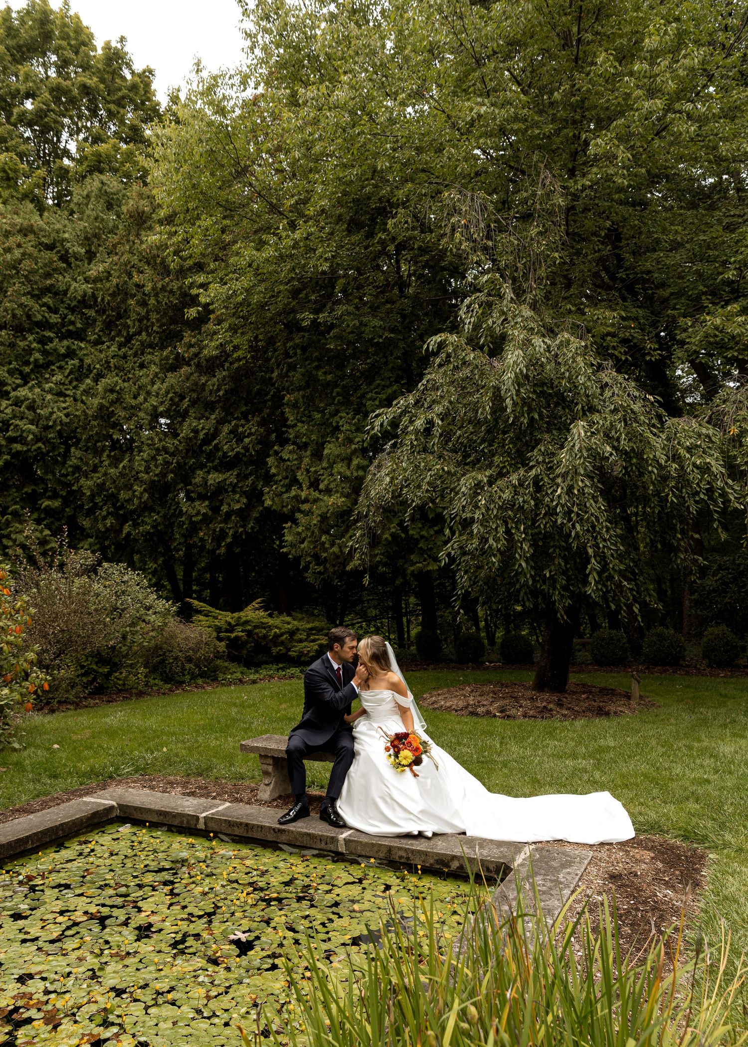 Bride and groom share an intimate moment near a garden pond with lily pads.
