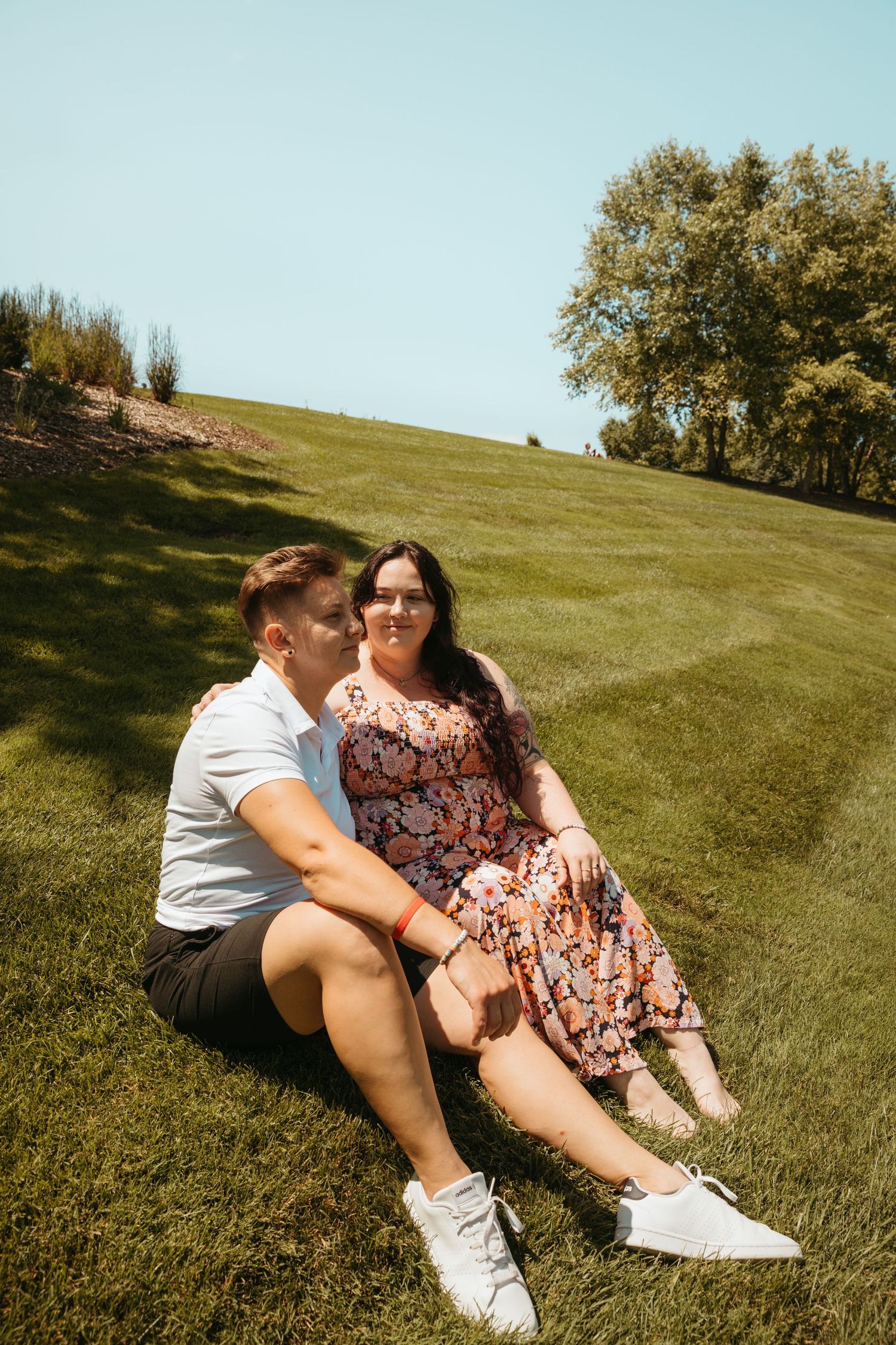 Two people share a tender moment while sitting on a green hillside under a bright blue sky.