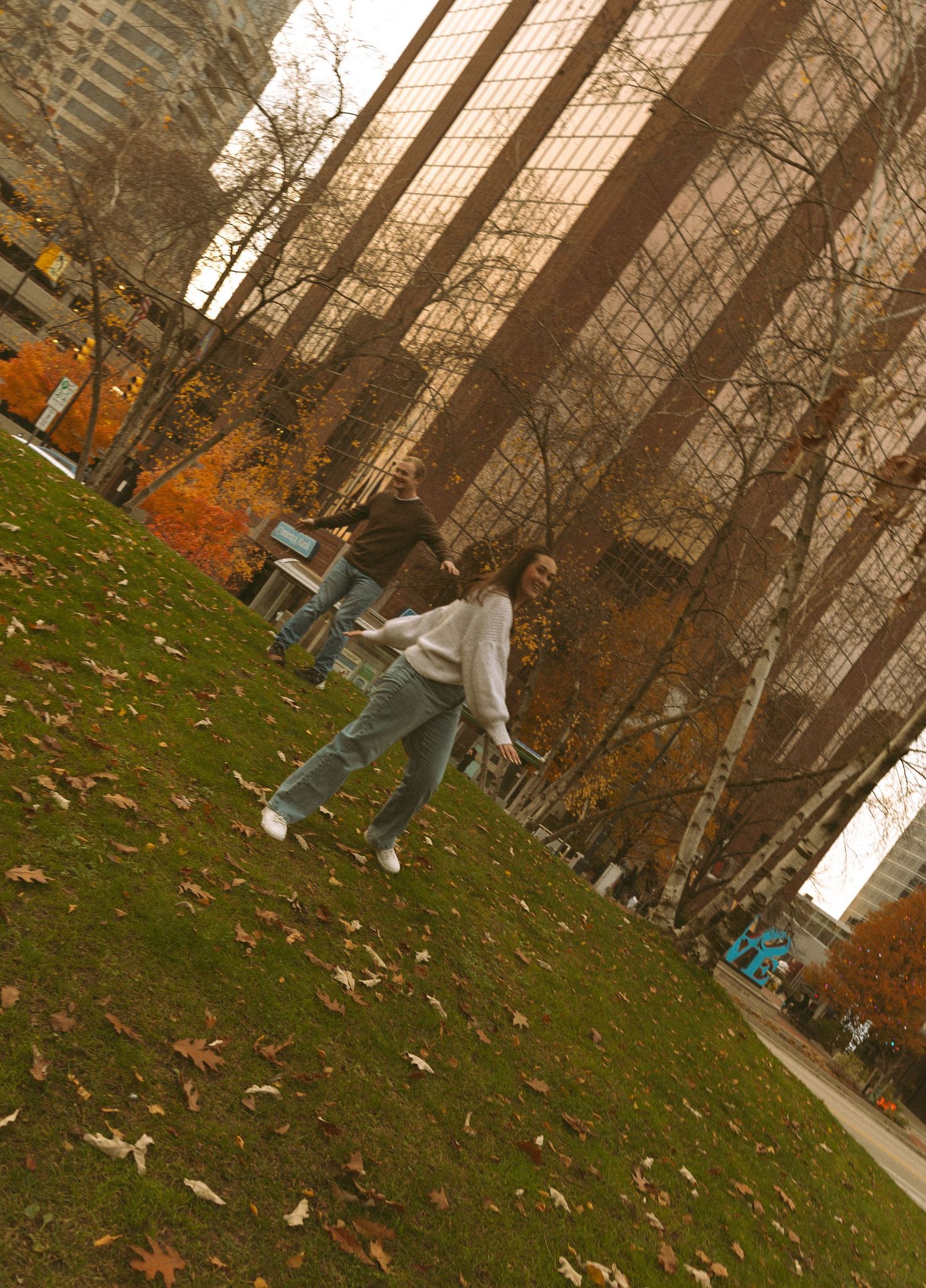 Dancing figures twirl on grass against a backdrop of autumn trees and modern architecture.