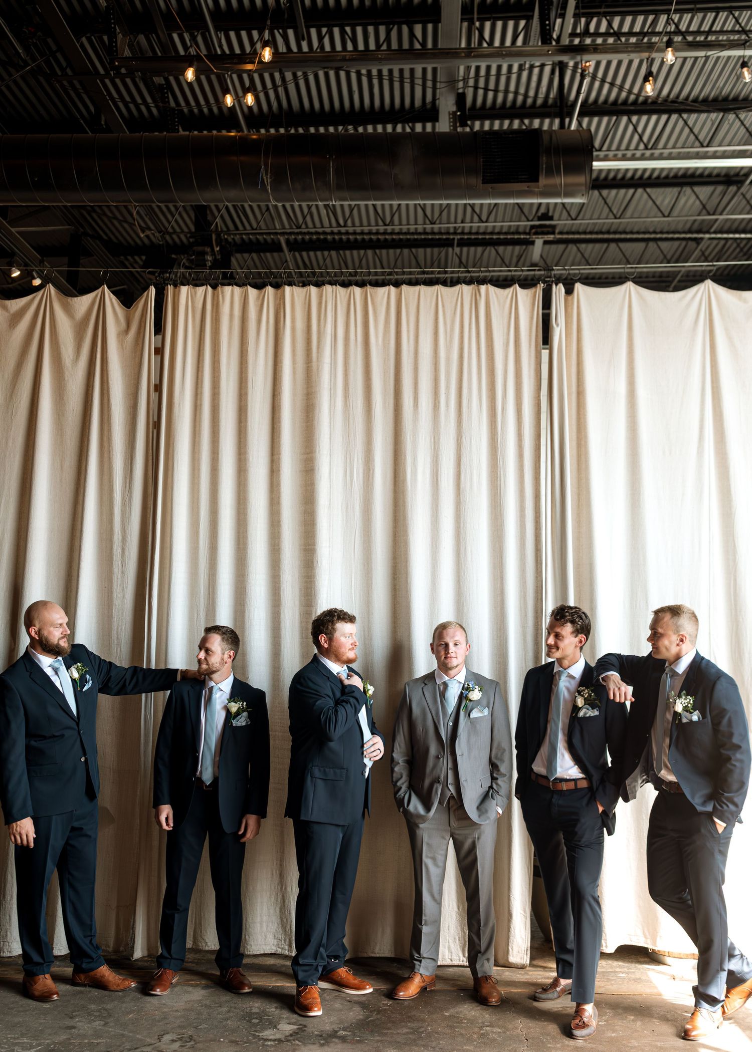 Groomsmen in suits pose together against white curtain backdrop in industrial venue.