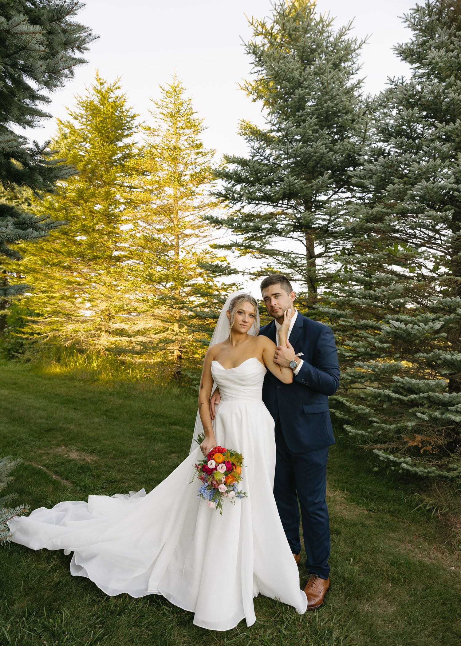 A wedding couple poses in front of evergreen trees with golden autumn foliage at sunset.