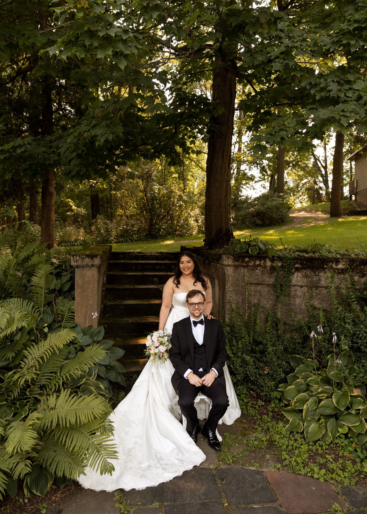 Wedding couple poses near stone walls and lush garden foliage.