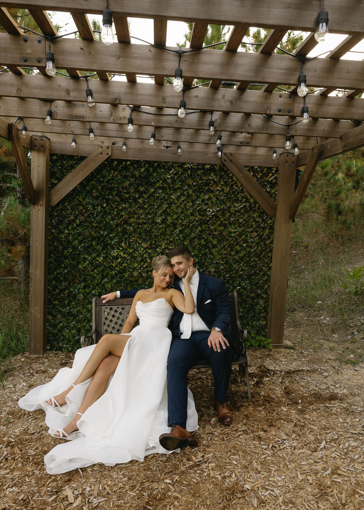 A couple shares intimate moments under a wooden pergola with string lights and ivy.