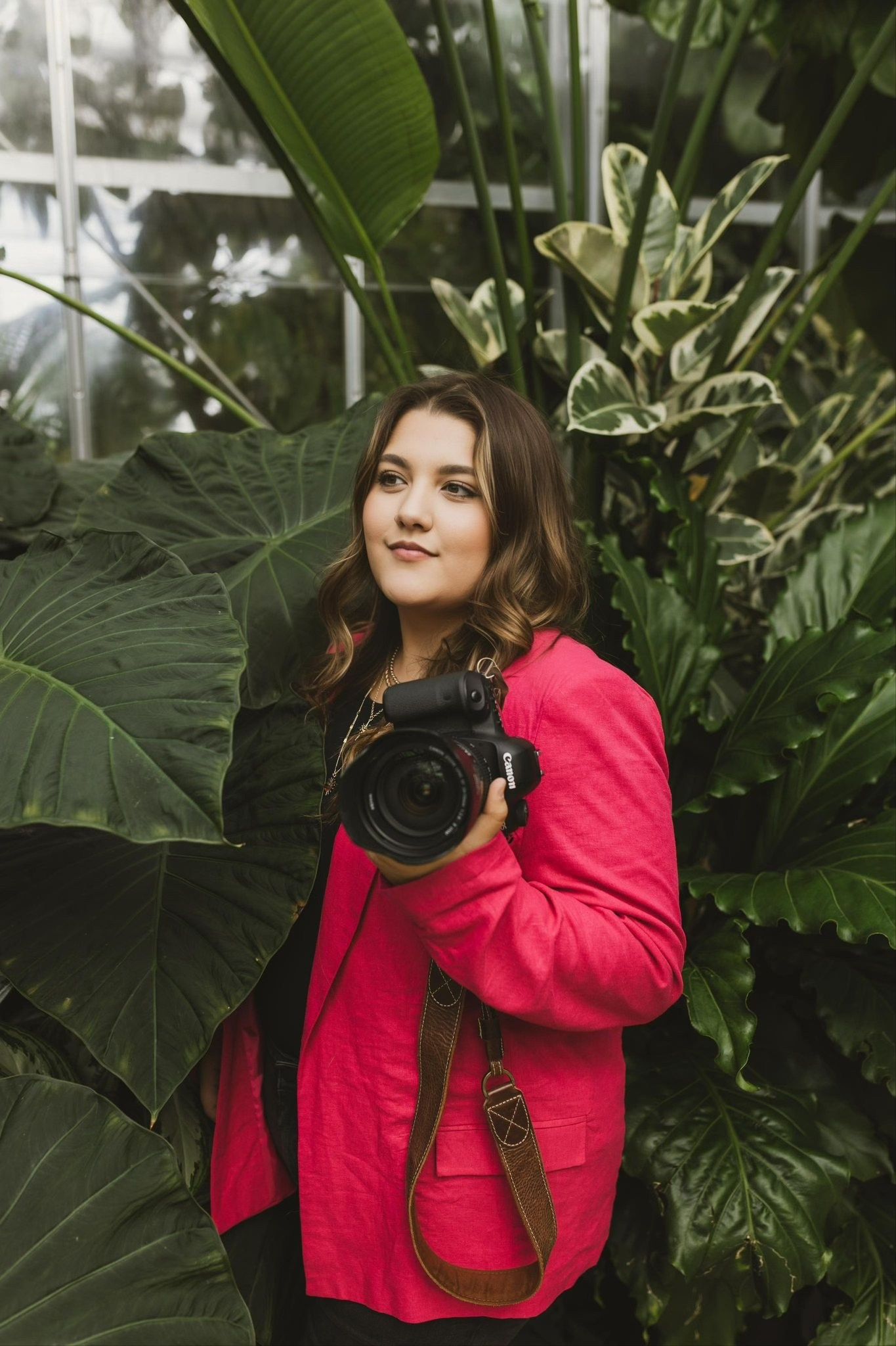 Professional photographer in bright red jacket poses with camera among tropical plants.