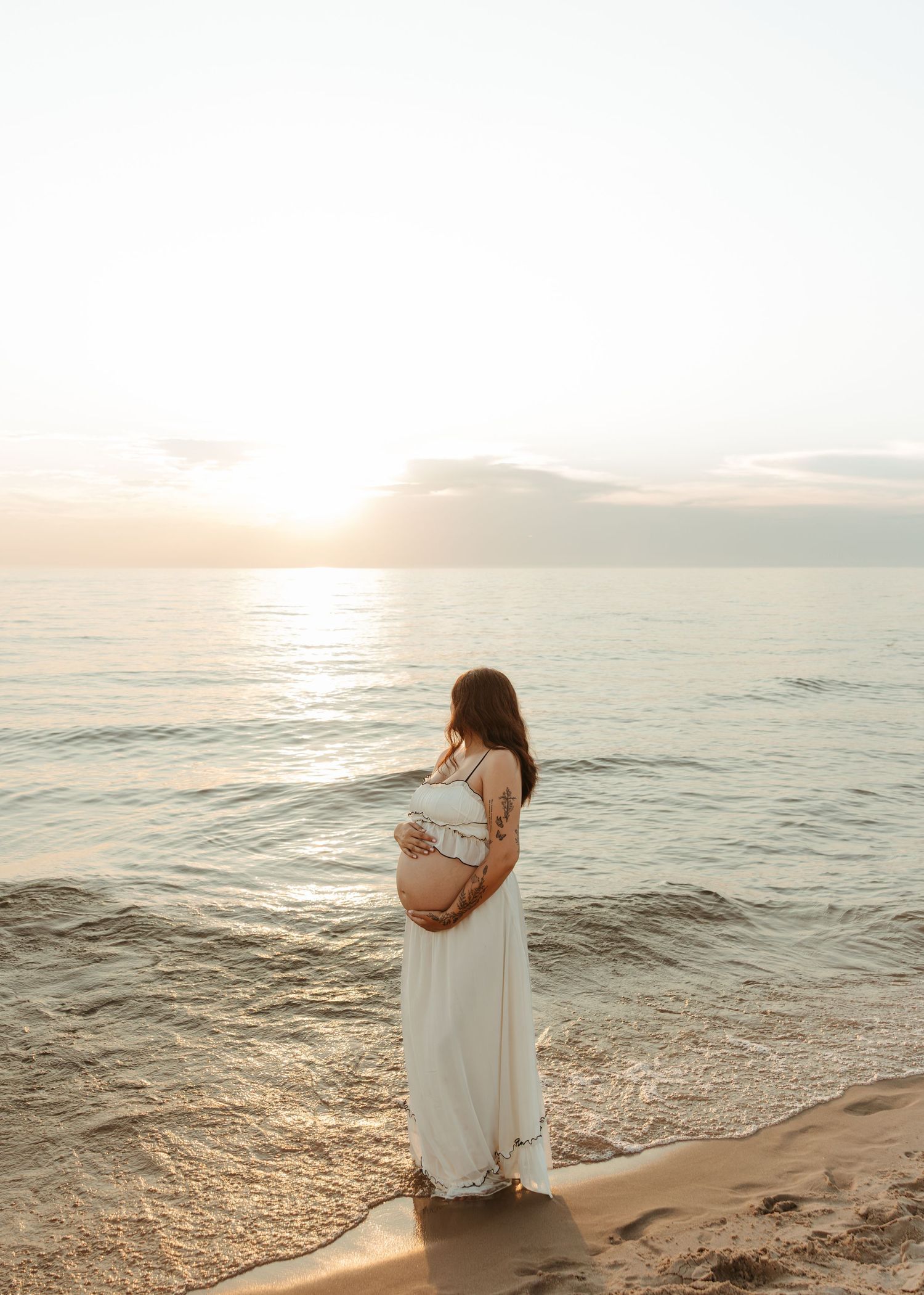 Expectant mother in a white dress stands contemplatively at the ocean's edge during sunset.
