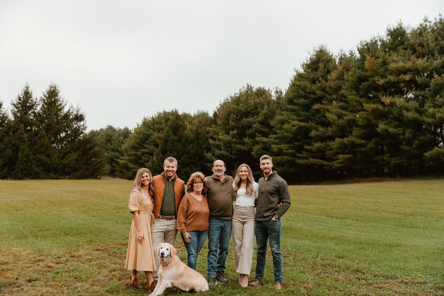 People and dog posing together on a grassy slope with pine trees in the background.