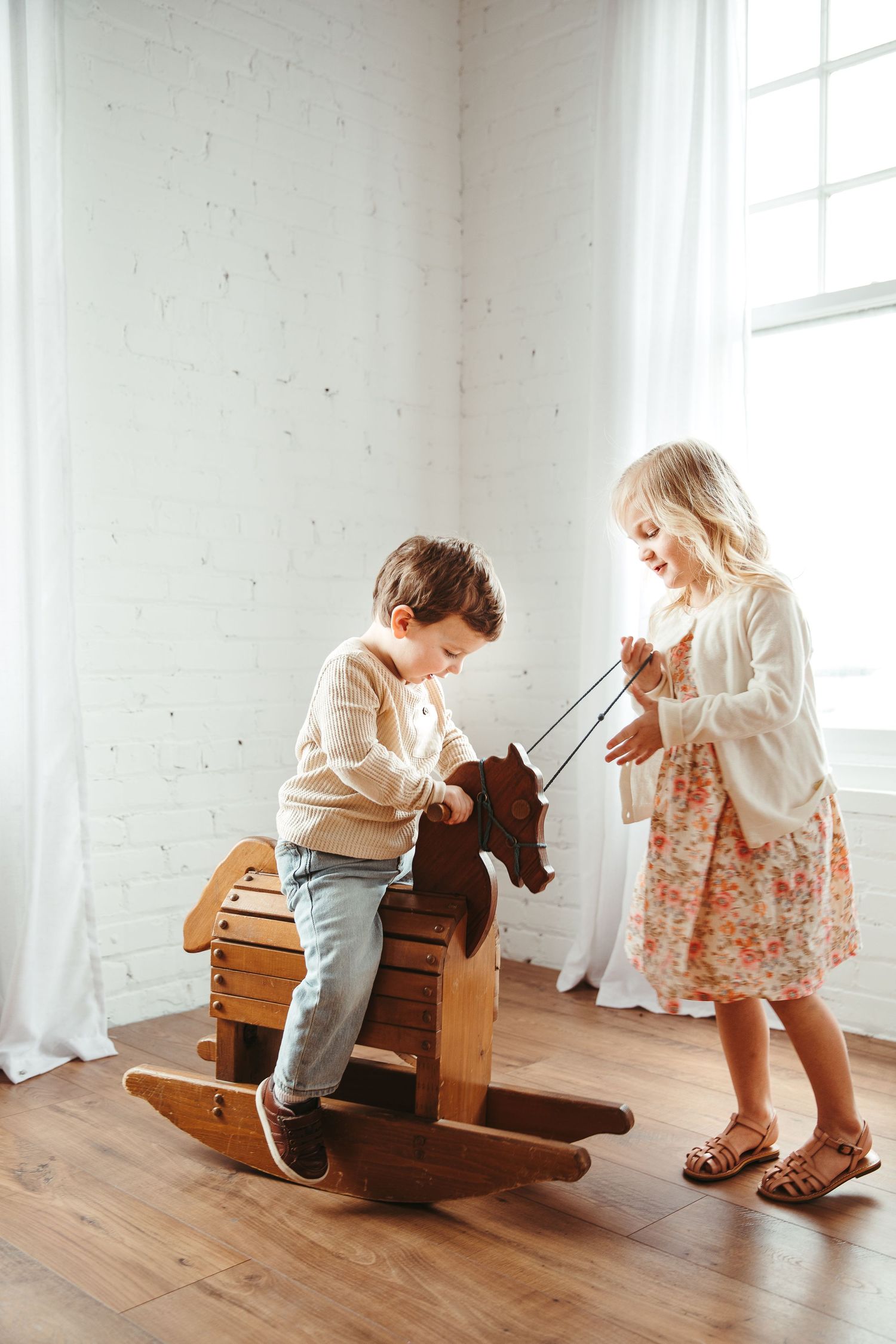 Children playing with wooden rocking horse in sunlit white room.