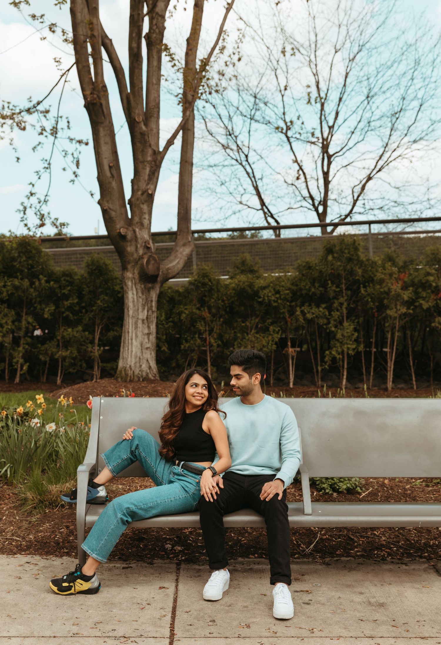 A couple relaxes casually on a concrete bench near trees on a sunny day.
