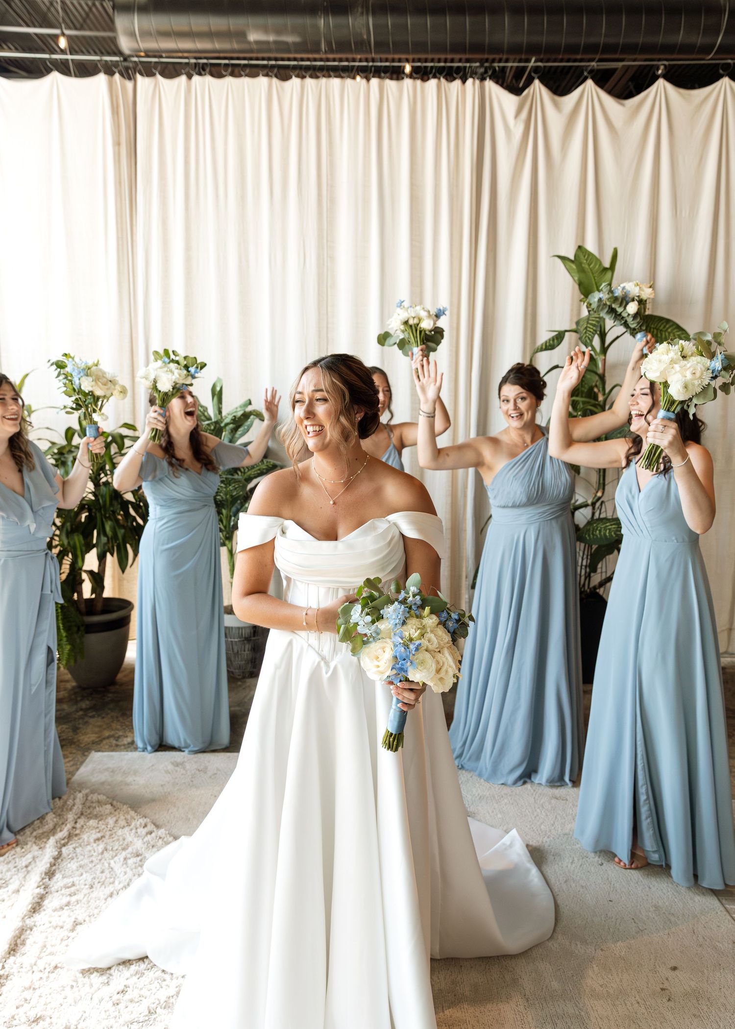 Bridal party in flowing blue gowns celebrates joyfully with bouquets against white curtain backdrop.