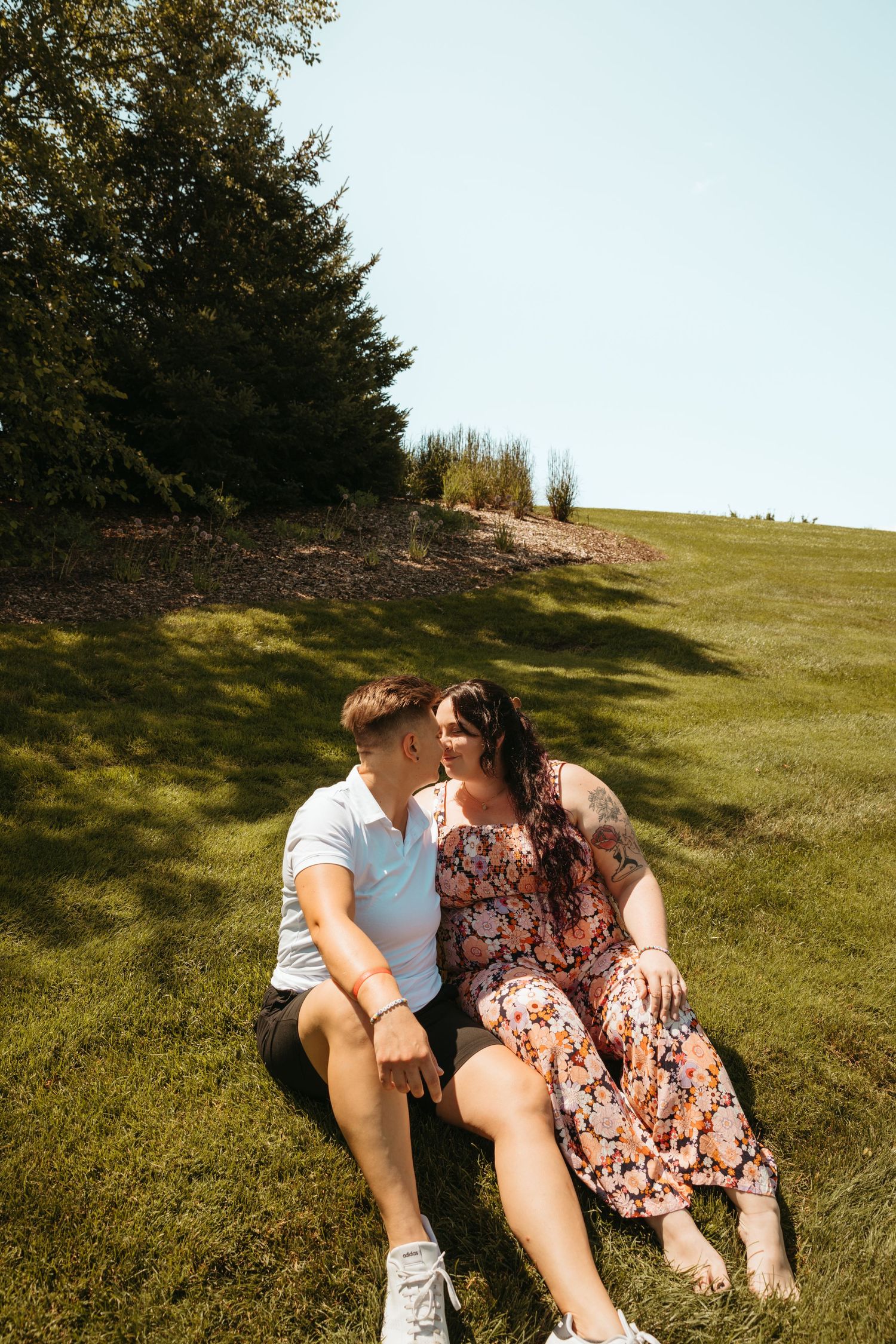 Two people share an intimate moment while sitting on a sunlit grassy slope with trees in the background.