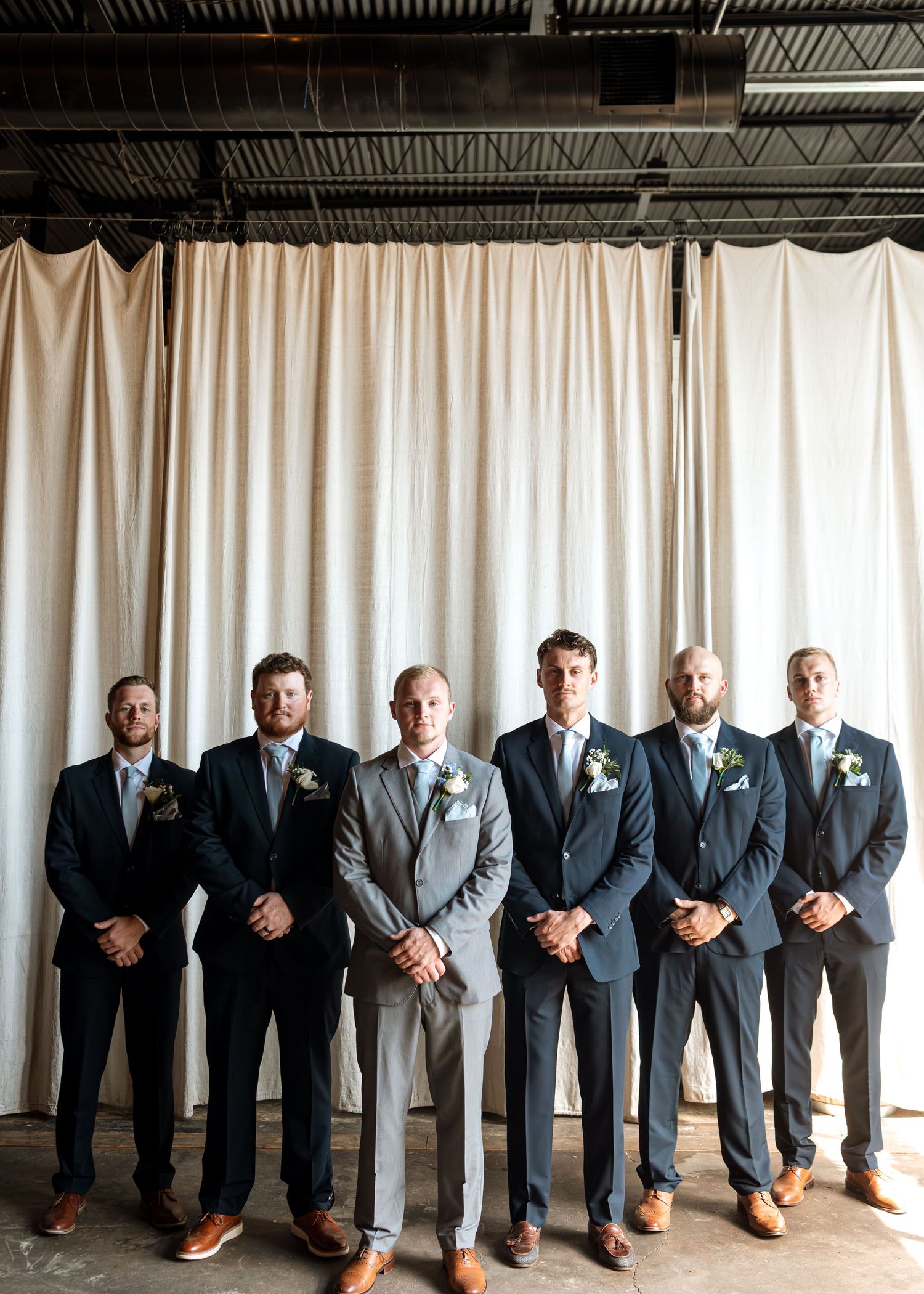 Groomsmen in coordinated suits and ties pose together against a white curtain backdrop.