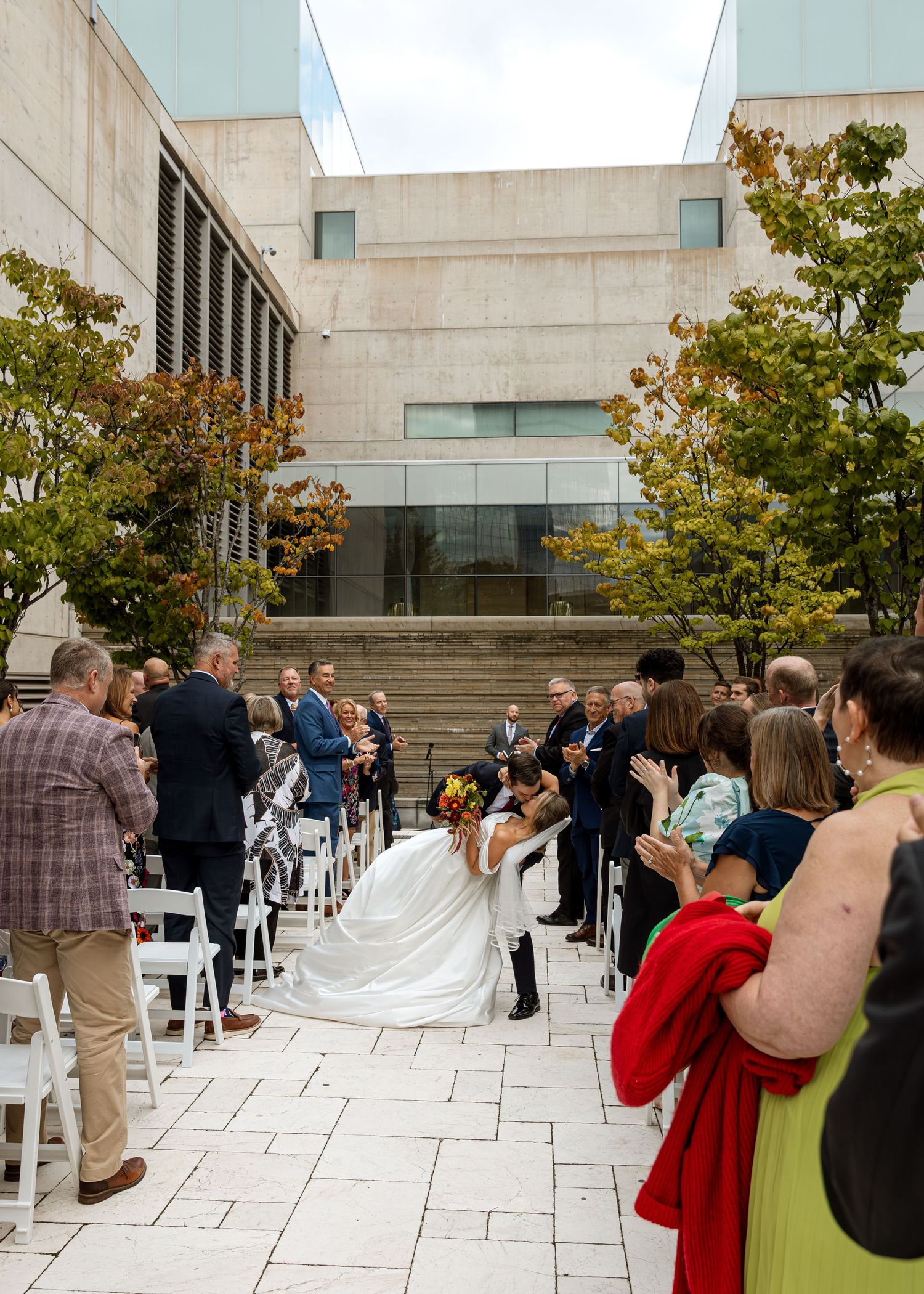 Wedding guests take photos of the newlyweds on a modern stone courtyard with autumn trees.