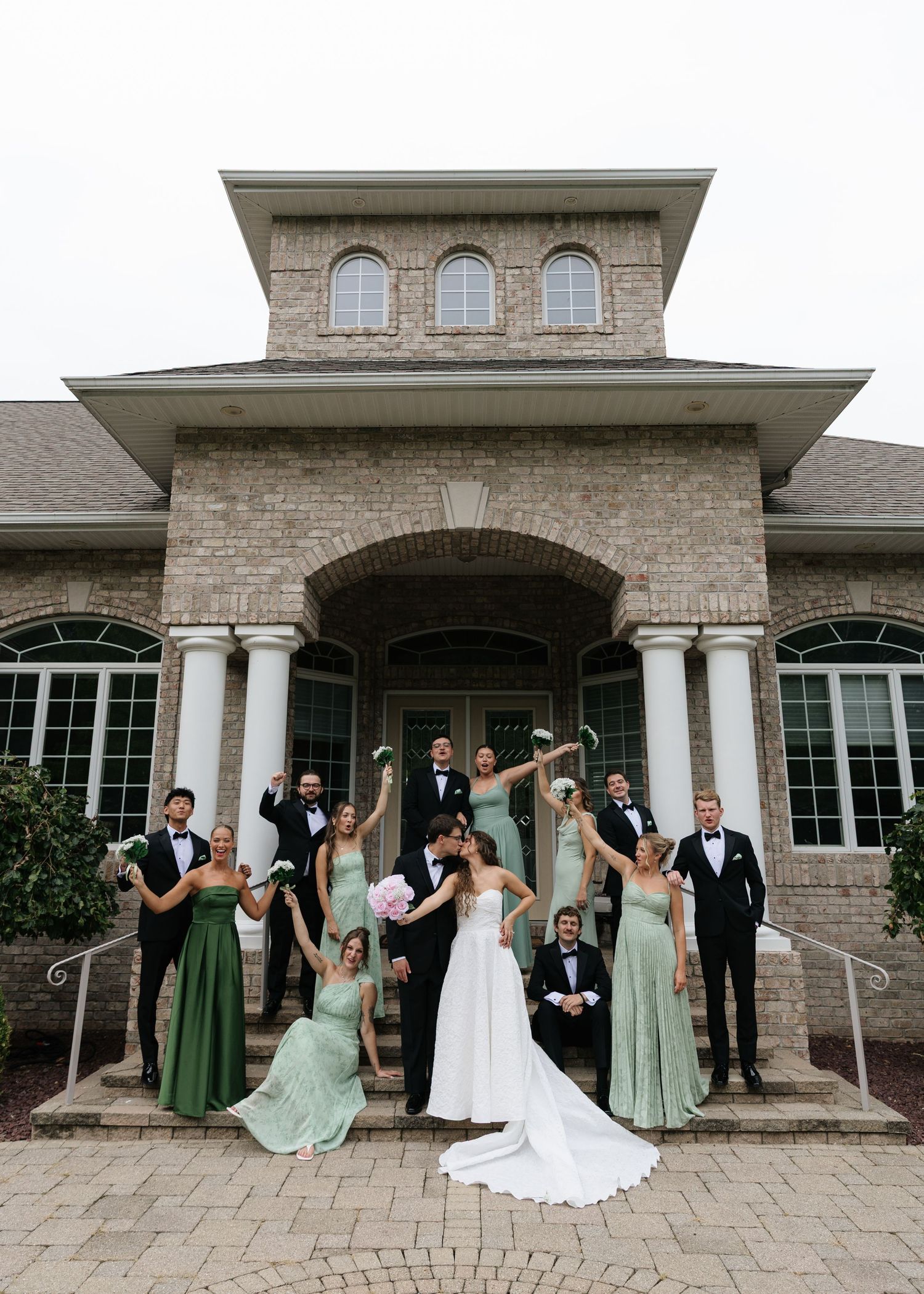 Wedding party poses outside brick venue, bride and groom kissing, bridesmaids in sage green gowns, groomsmen in black tuxedos.