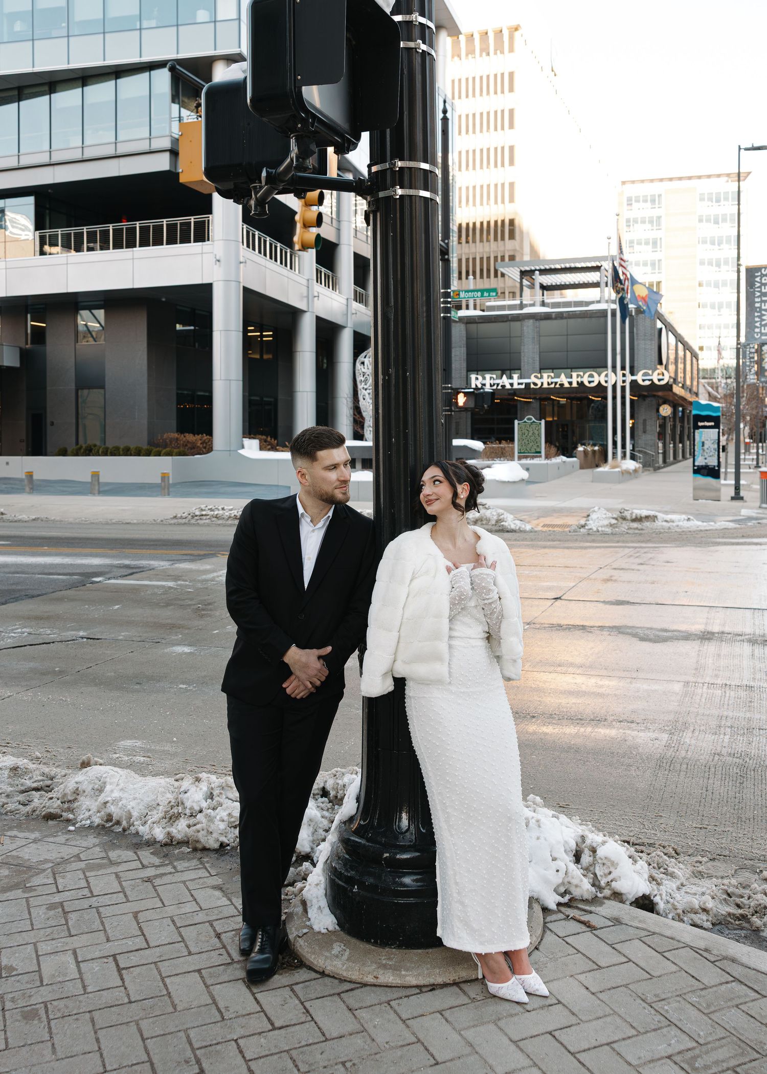 Couple in wedding attire posing by a lamppost on a snowy urban street corner, with city buildings in the background.