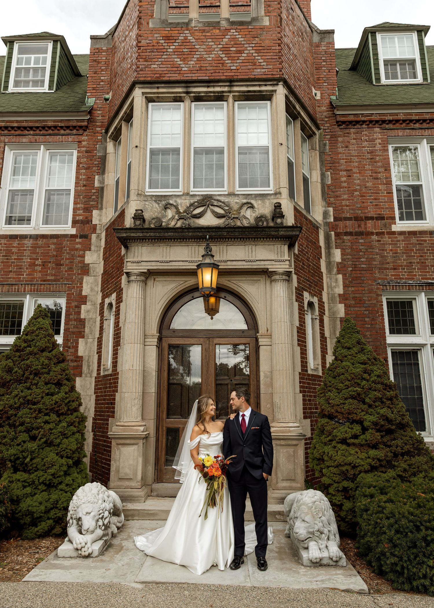 Newlyweds stand between decorative stone lions at a grand building entrance.
