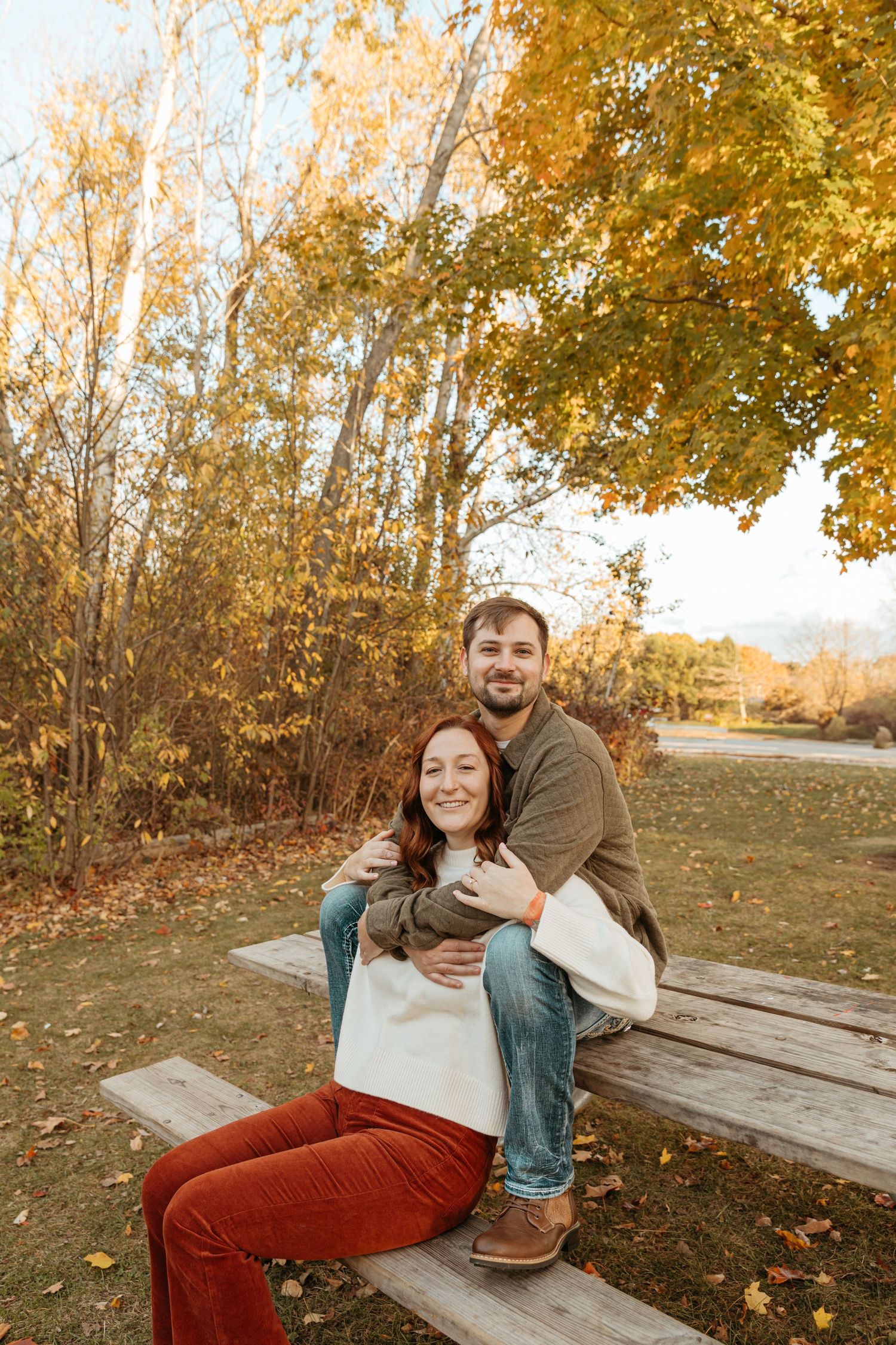 A couple shares a tender embrace while sitting on a wooden bench surrounded by golden autumn foliage.