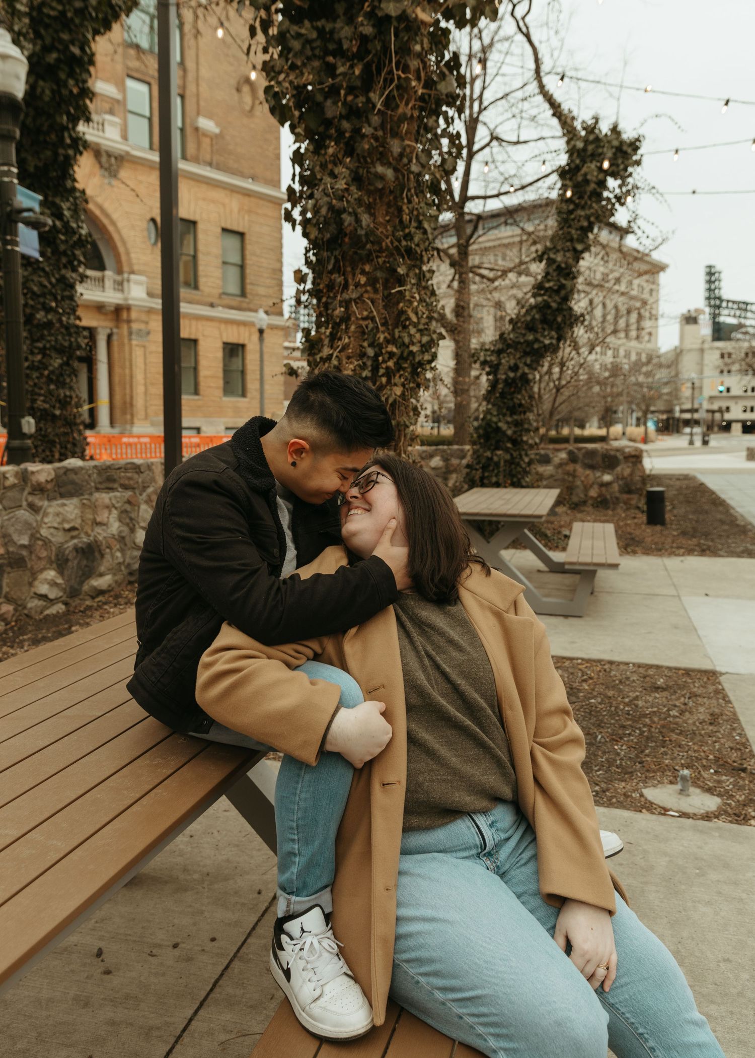 Couple sharing tender moment on urban wooden bench surrounded by ivy-covered buildings.