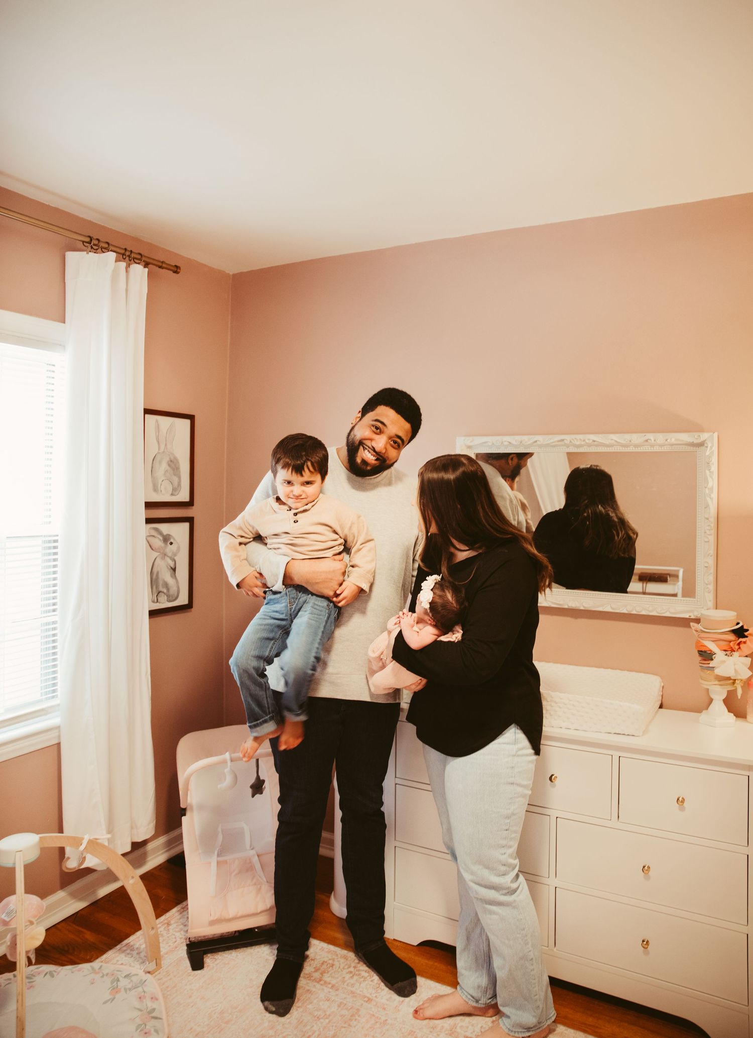 A family shares playful moments together in a bedroom with pink walls and white furniture.