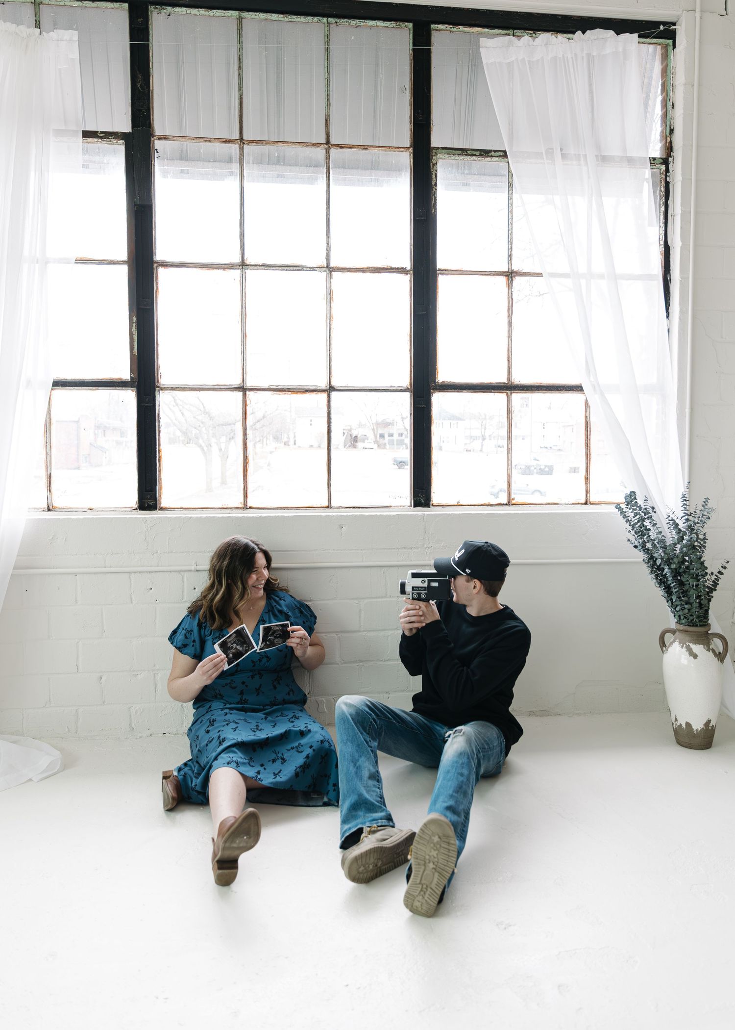 Couple sitting on white floor by large windows, woman holding ultrasound photos while man films her with vintage camera.
