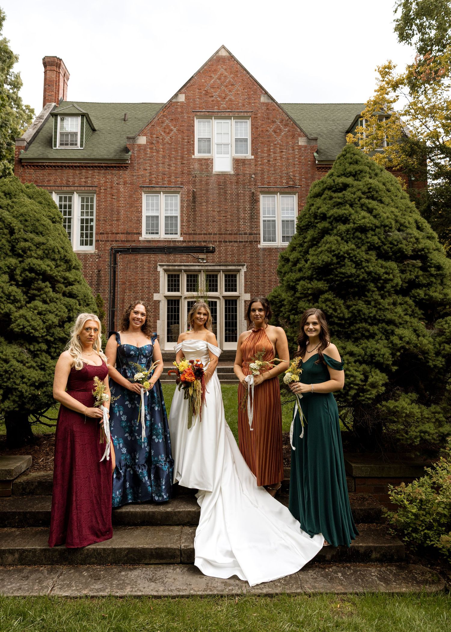Bridesmaids in burgundy and green dresses pose with a bride in white outside a brick mansion.