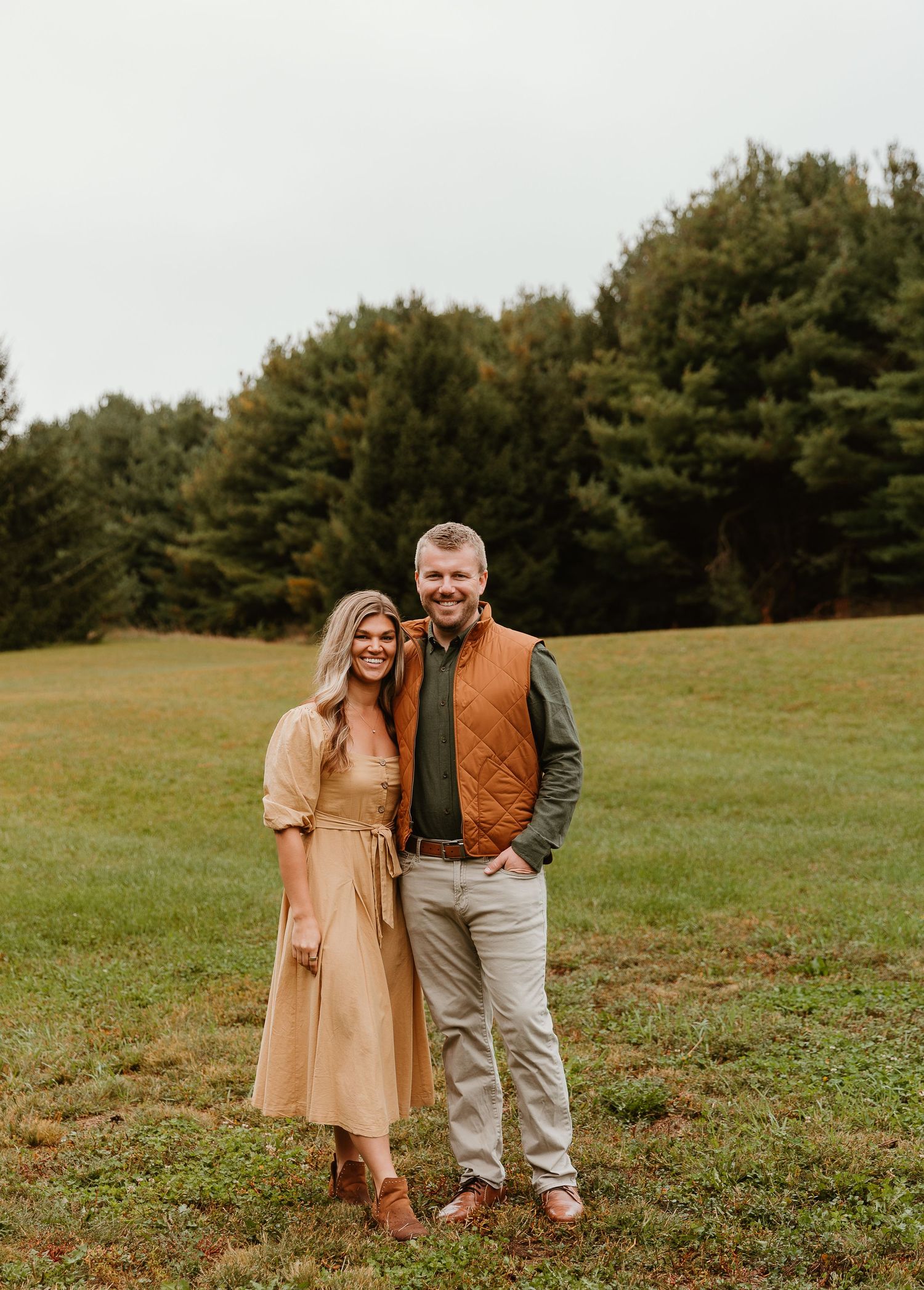 A couple poses together in a peaceful meadow surrounded by autumn trees.