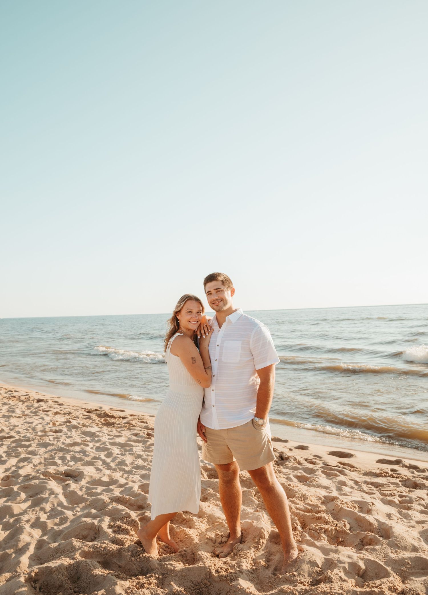 Couple in white outfits sharing a romantic moment on a sunny beach at sunset.