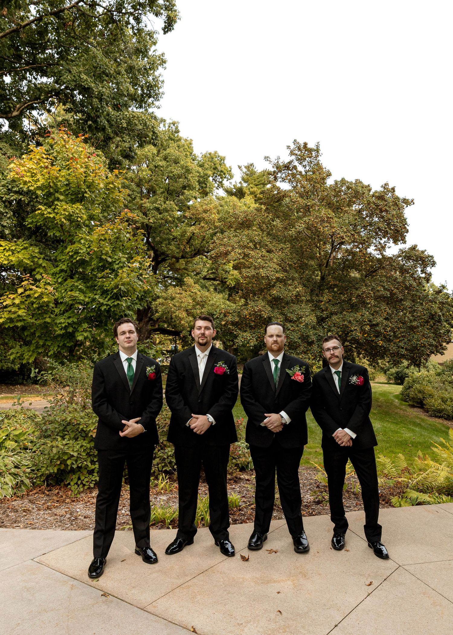 Groomsmen in black suits stand together on a paved pathway surrounded by autumn trees.