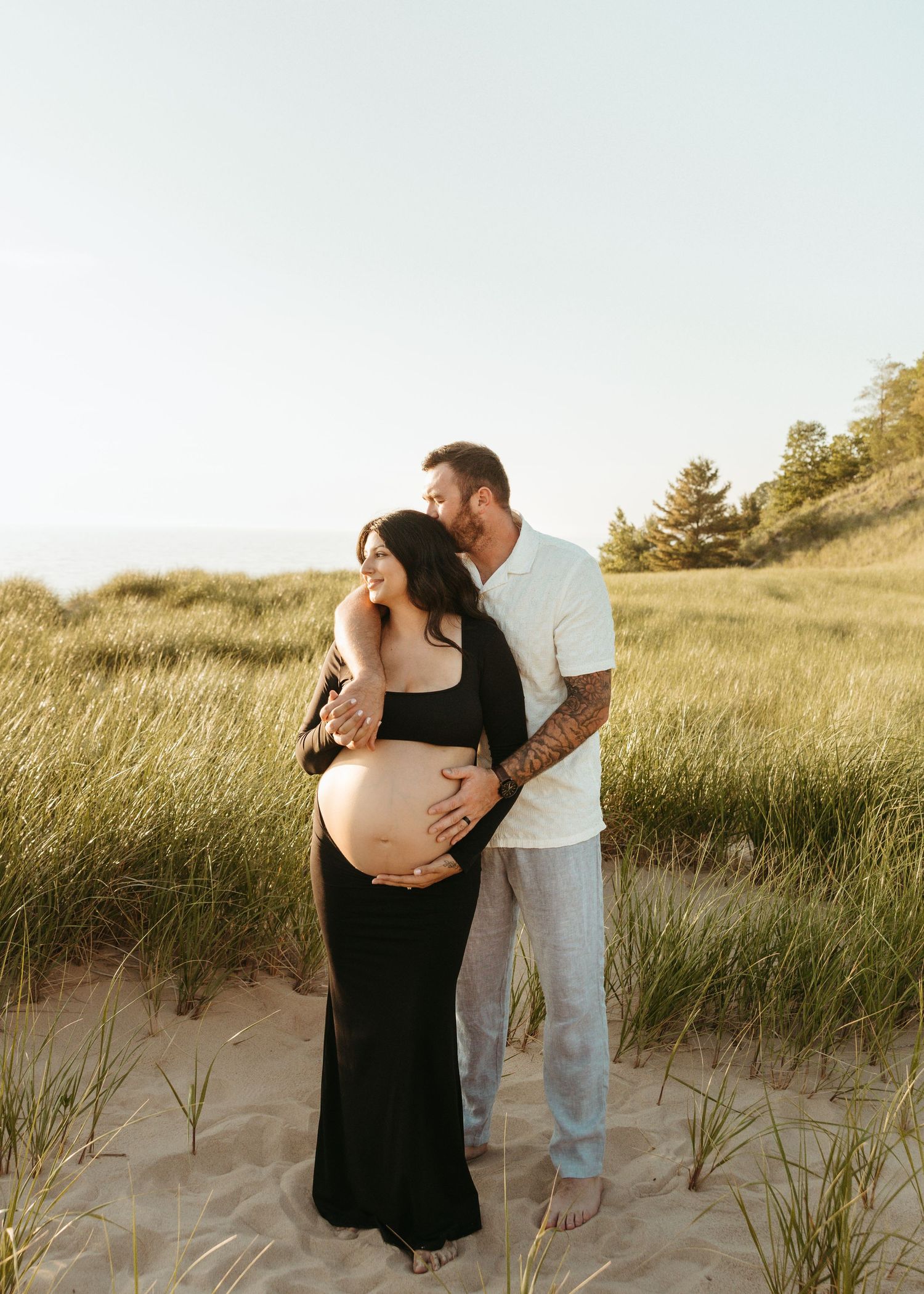 A maternity photoshoot featuring a couple standing among beach grass at dusk.