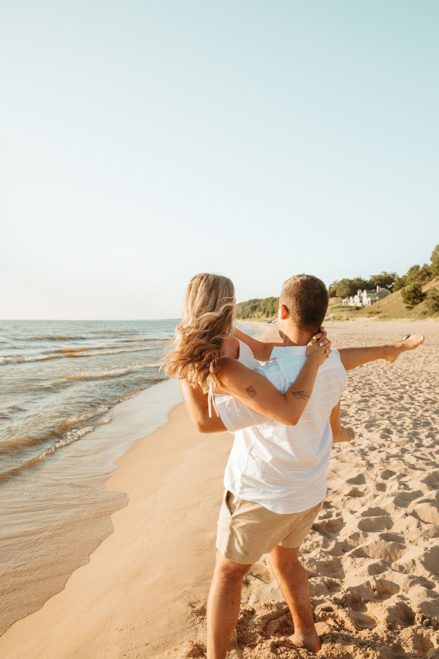 Playful couple in white outfits dancing on beach at sunset.