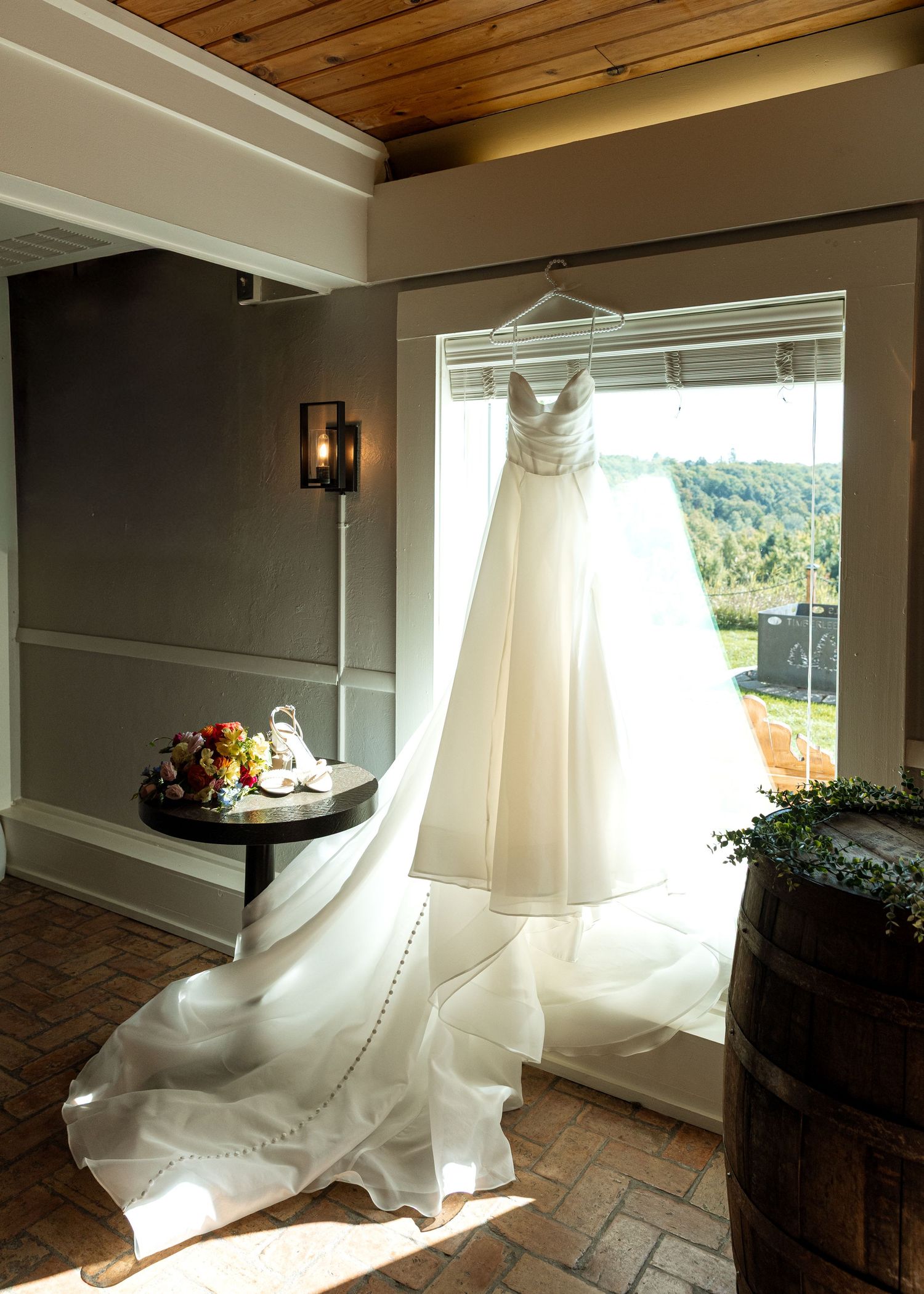 White wedding dress hanging in sunlit room with wooden ceiling and tile floor.