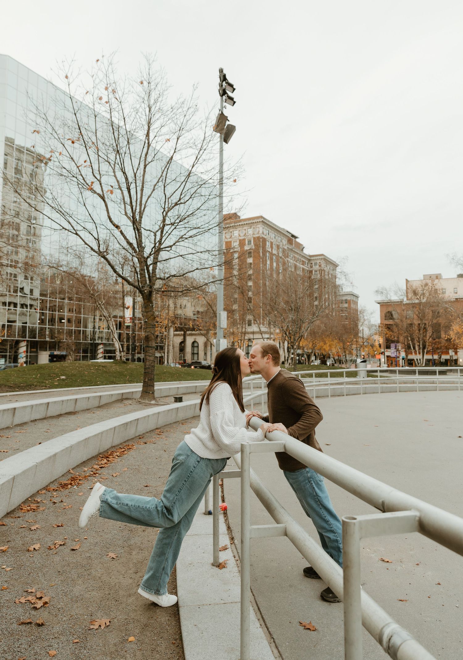 A couple shares a romantic moment leaning over concrete barriers in an urban setting during autumn.