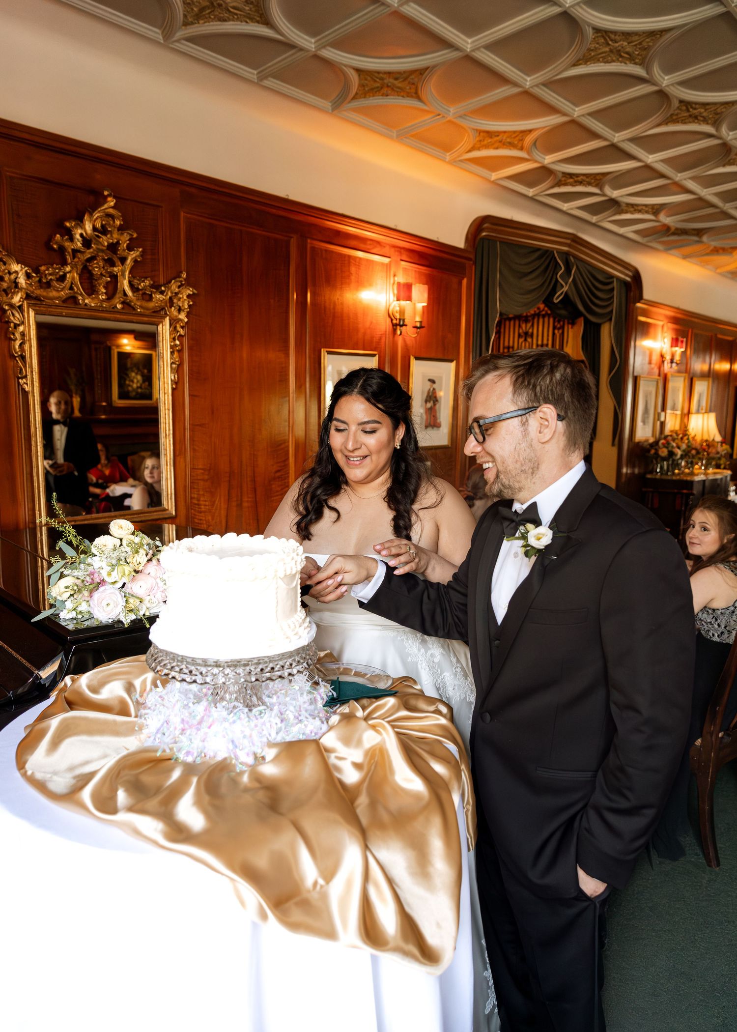 A wedding couple cuts their elegant white cake displayed on a gold satin tablecloth in an ornate wood-paneled room.
