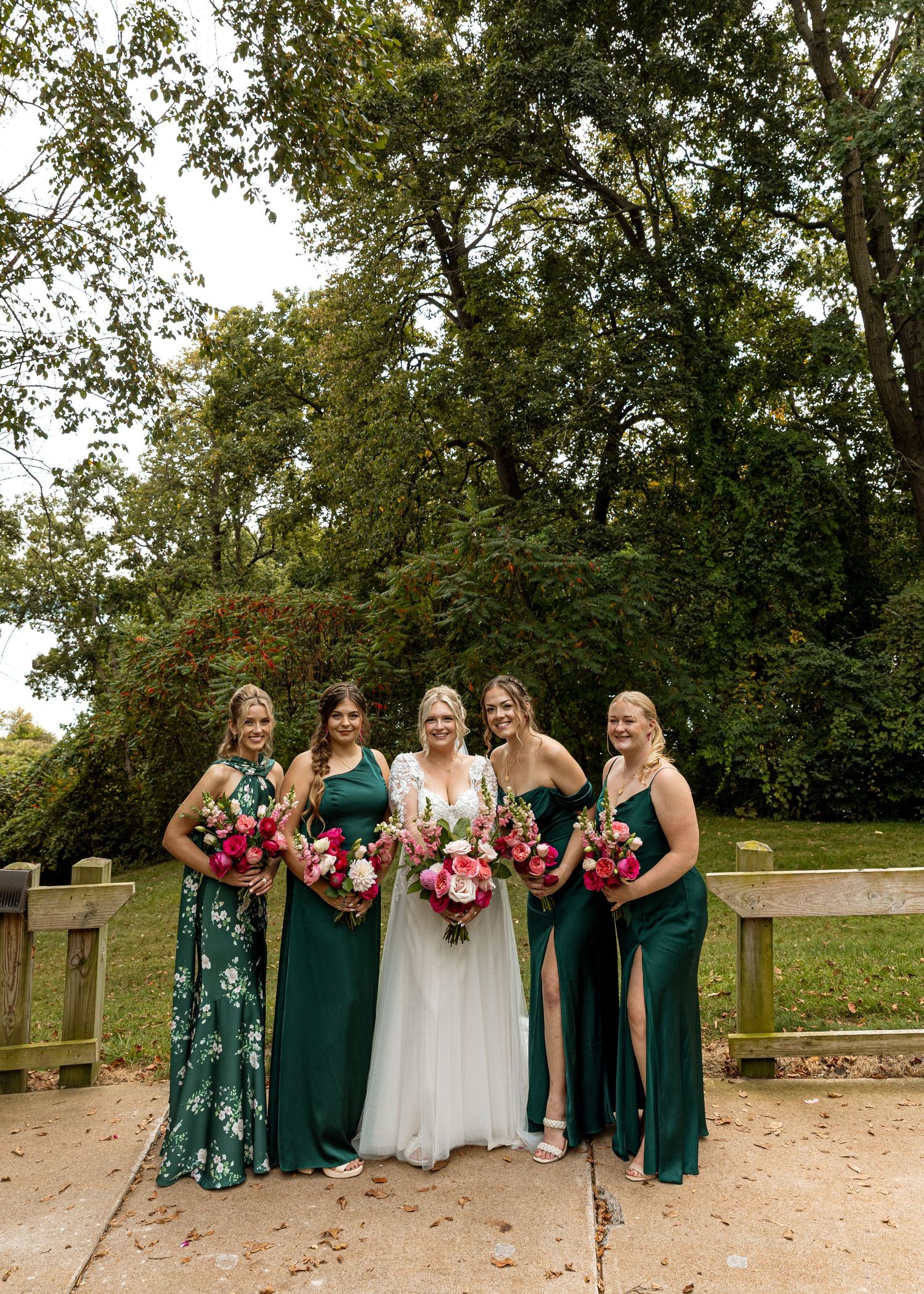Bridesmaids in elegant emerald dresses pose with bouquets on a garden path.