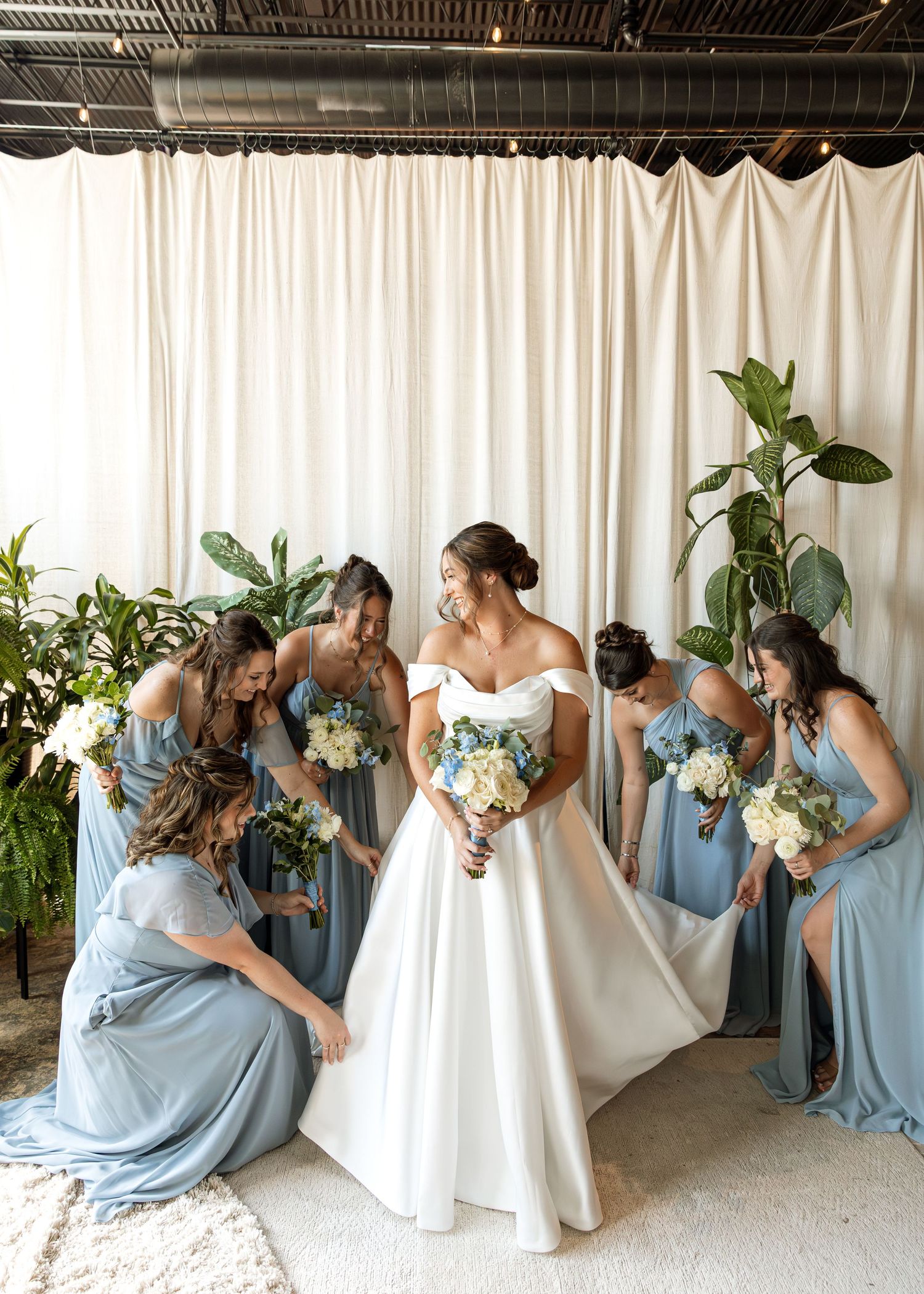 Wedding party in dusty blue dresses gathers around bride in elegant white gown.