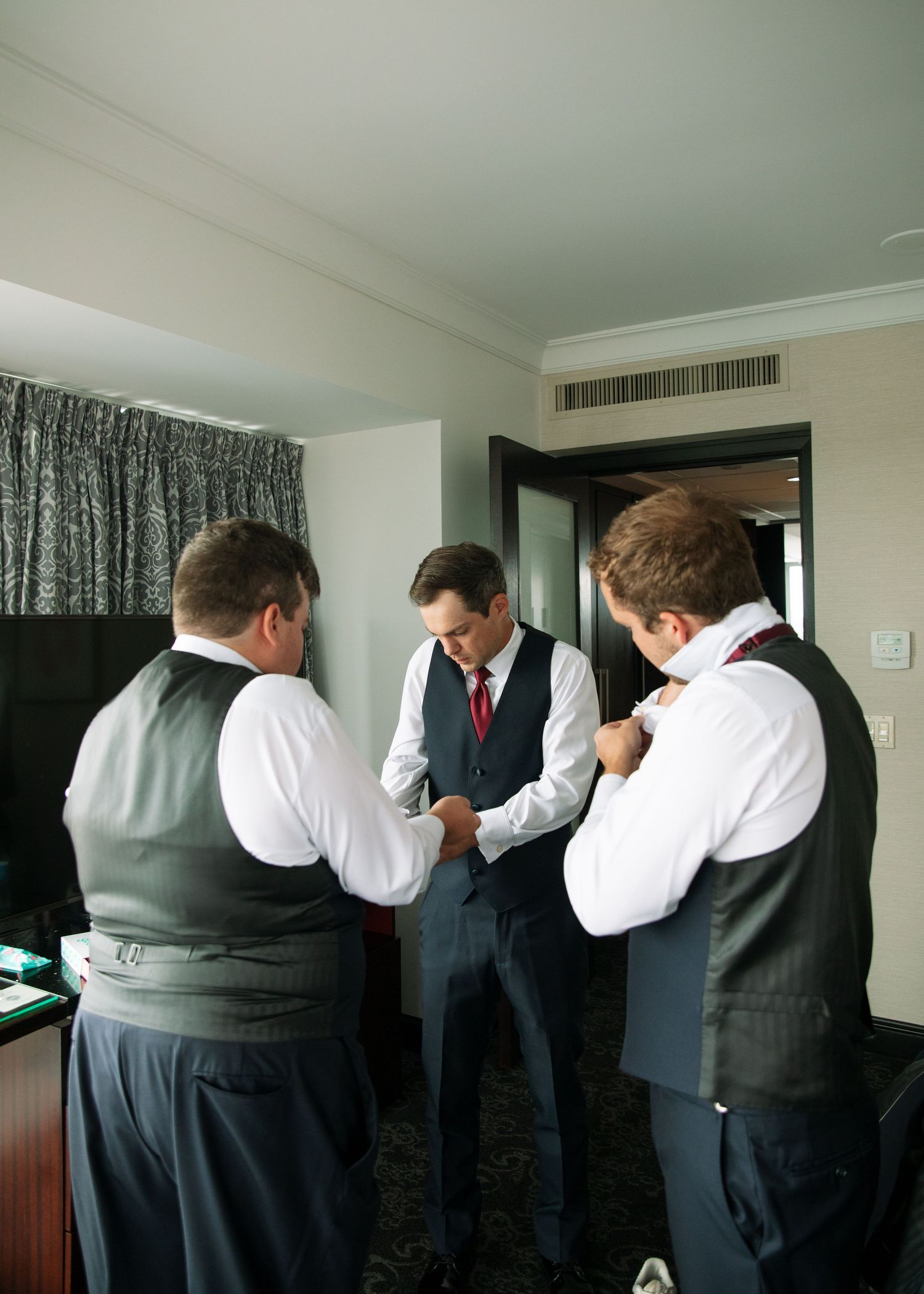 Groomsmen help each other get ready in a hotel room before the ceremony.