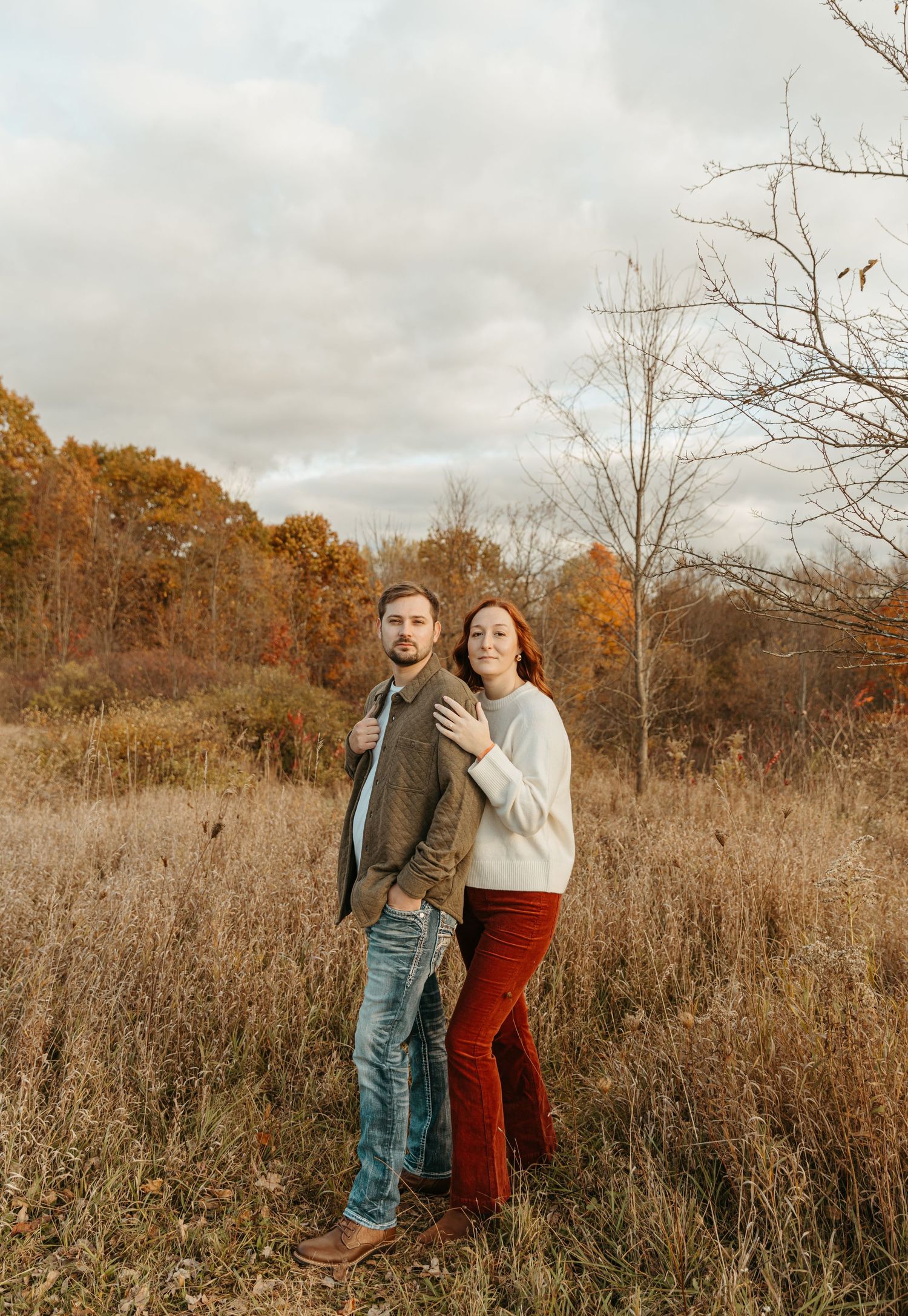 A couple stands together in a golden autumn field with tall grass and trees.