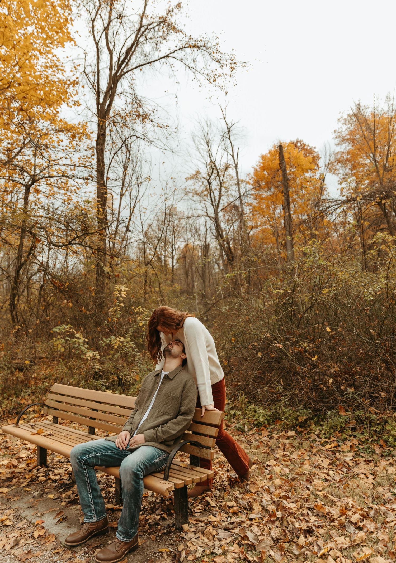 A couple shares a playful moment on a wooden bench amid fallen autumn leaves.