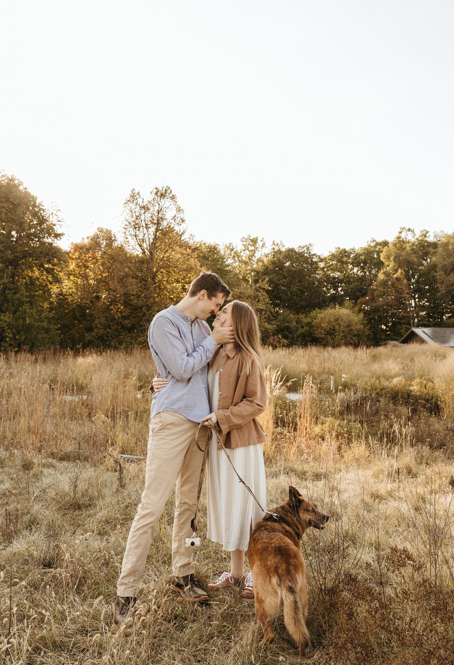 Romantic couple embracing in a golden field at sunset with their German Shepherd dog.