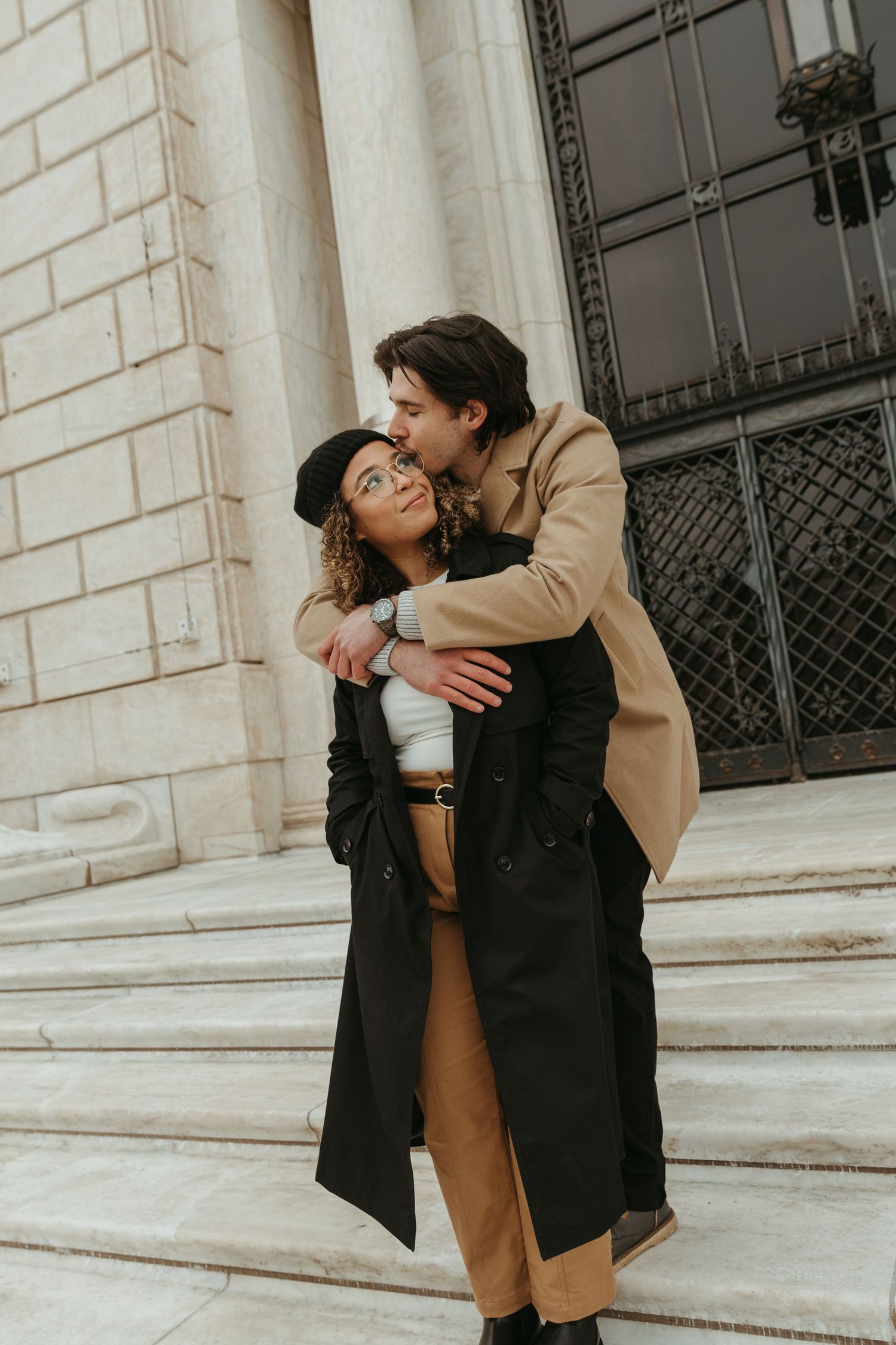 A romantic couple embraces on white stone steps of a classical building.