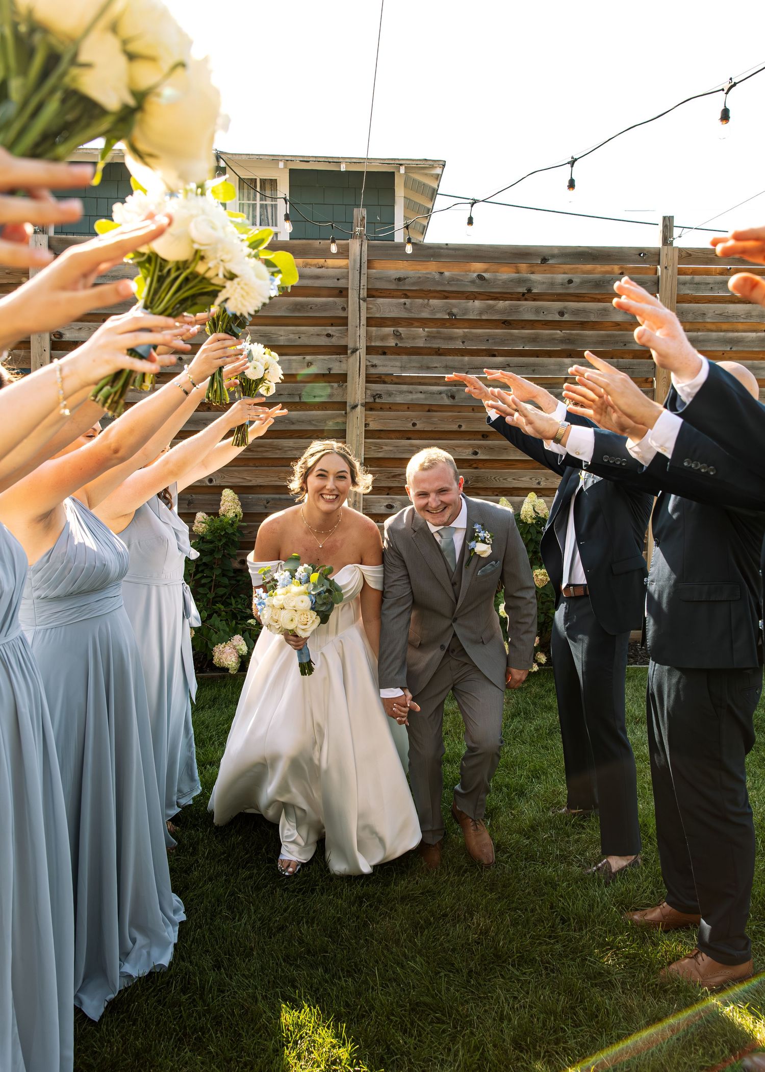 Wedding party celebrates with raised arms as couple walks under string lights in outdoor garden setting.