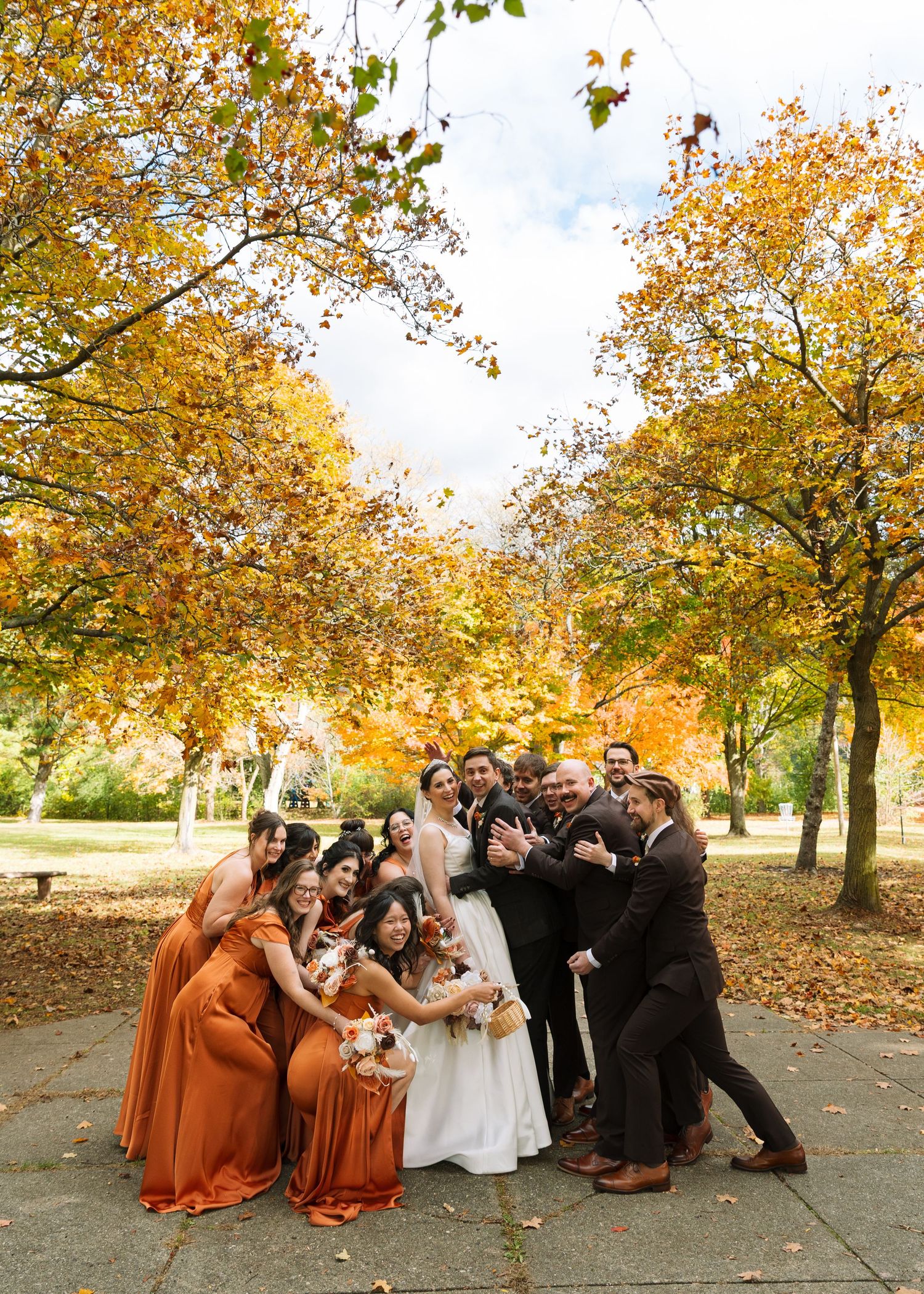 Wedding party celebrating outdoors among vibrant autumn trees, bridesmaids in rust gowns and groomsmen in brown suits surrounding the bride.