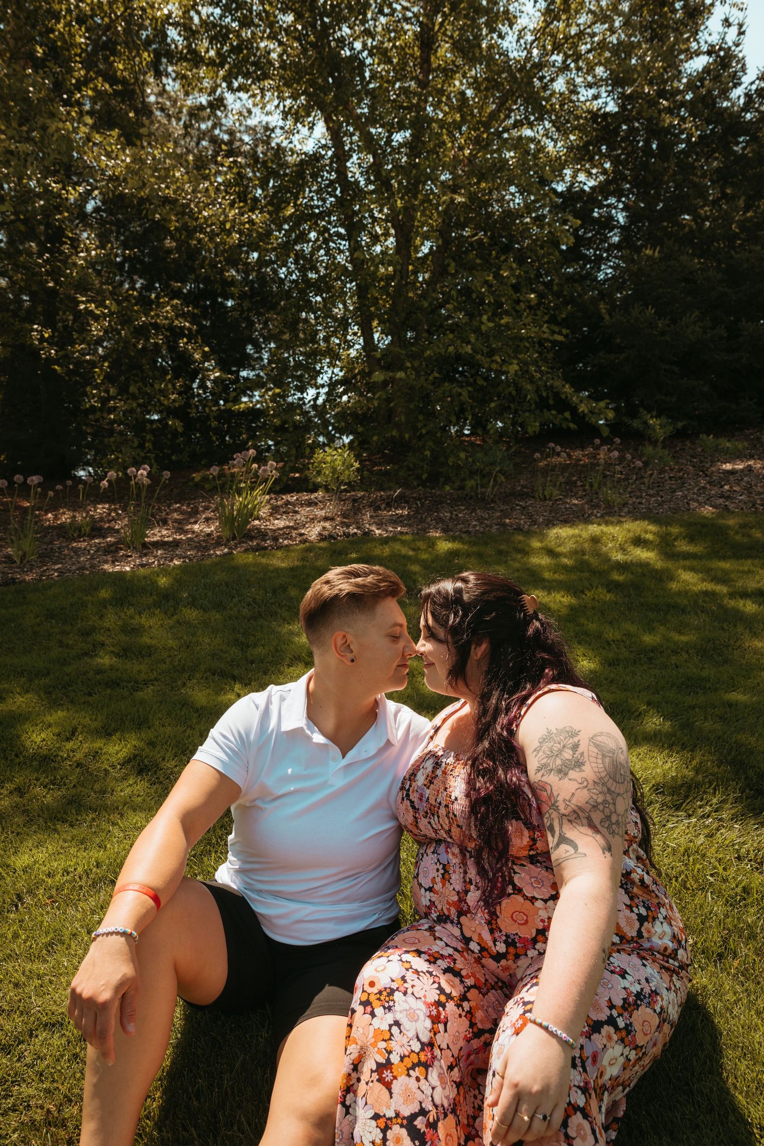 A couple relaxes on a grassy hill during a warm day, surrounded by trees and natural scenery.