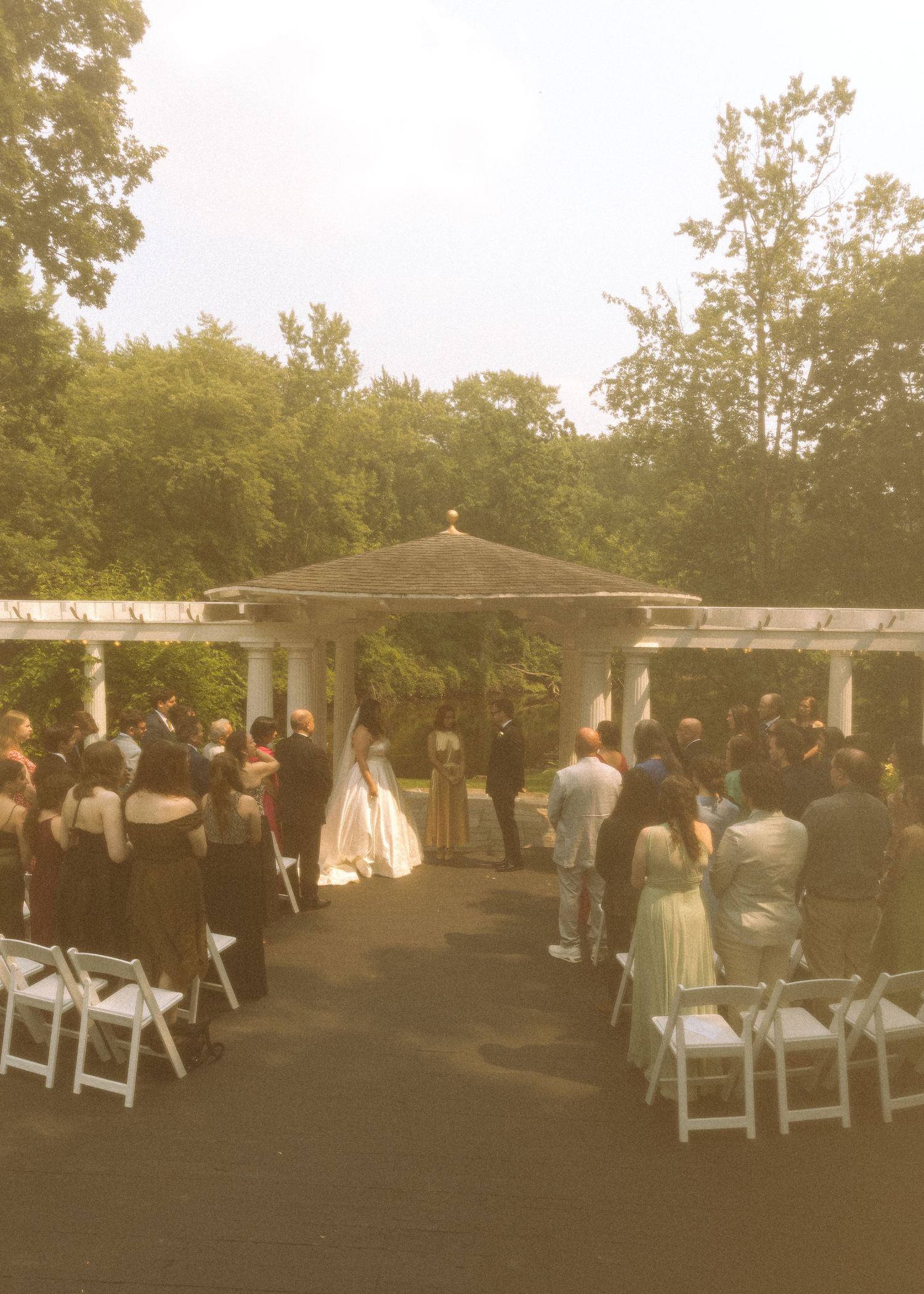 Outdoor wedding ceremony setup with white chairs and guests gathering under trees.