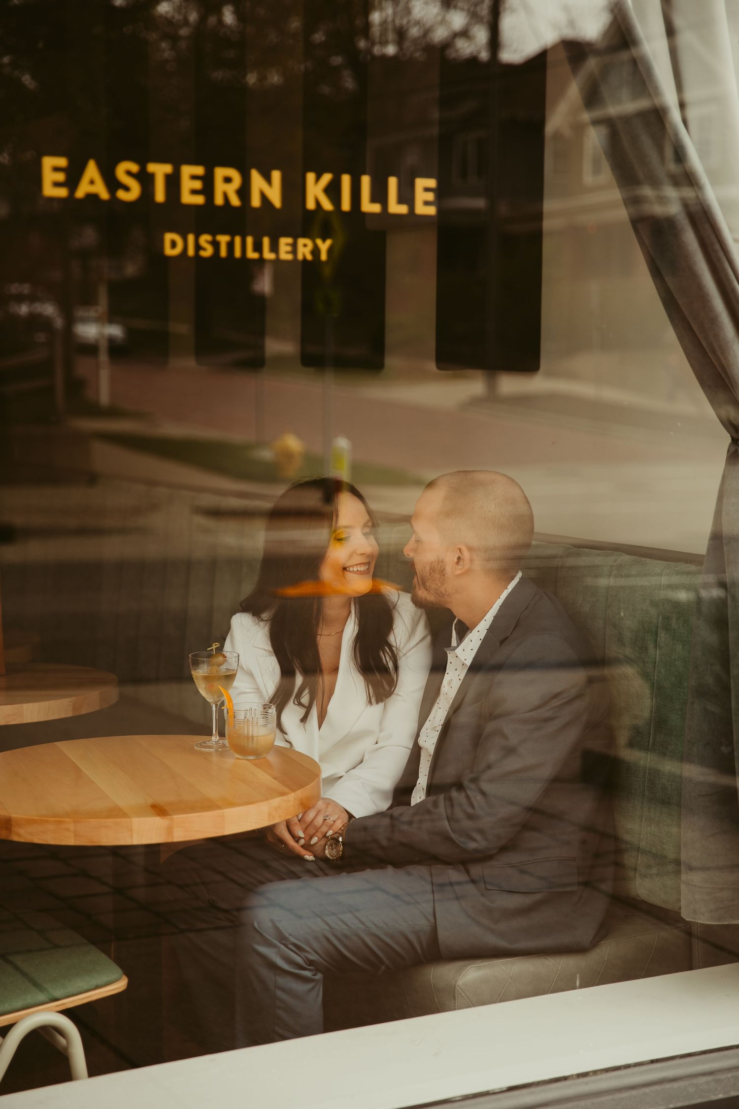 Couple sits at table inside Eastern Kille Distillery viewed through window.