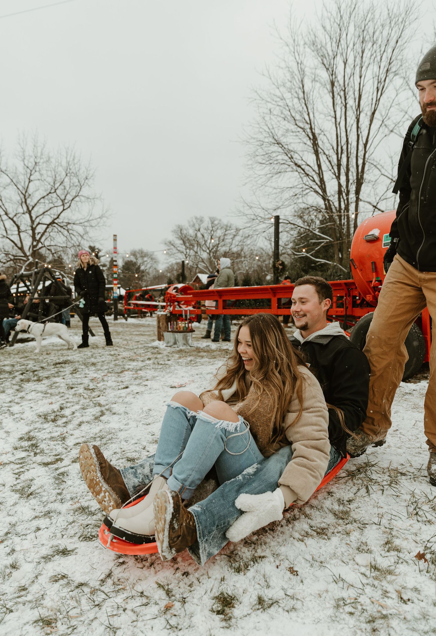 Couple playfully sledding together in snowy winter landscape with red barn in background.